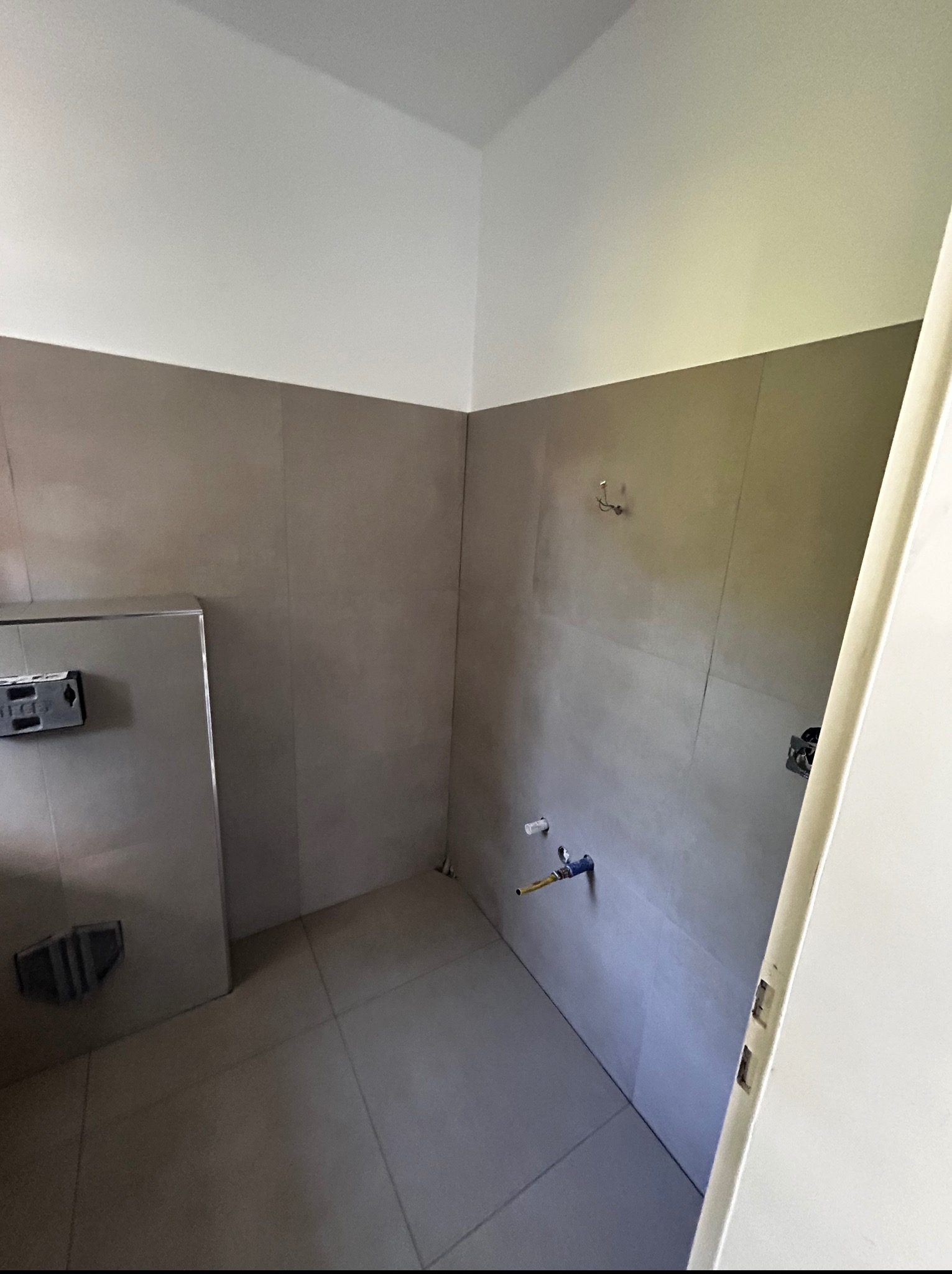 Wnętrze łazienki w trakcie remontu. Widoczne płytki na ścianach, podłączenia wodne i fragment drzwi. Przygotowanie do dalszych prac wykończeniowych.