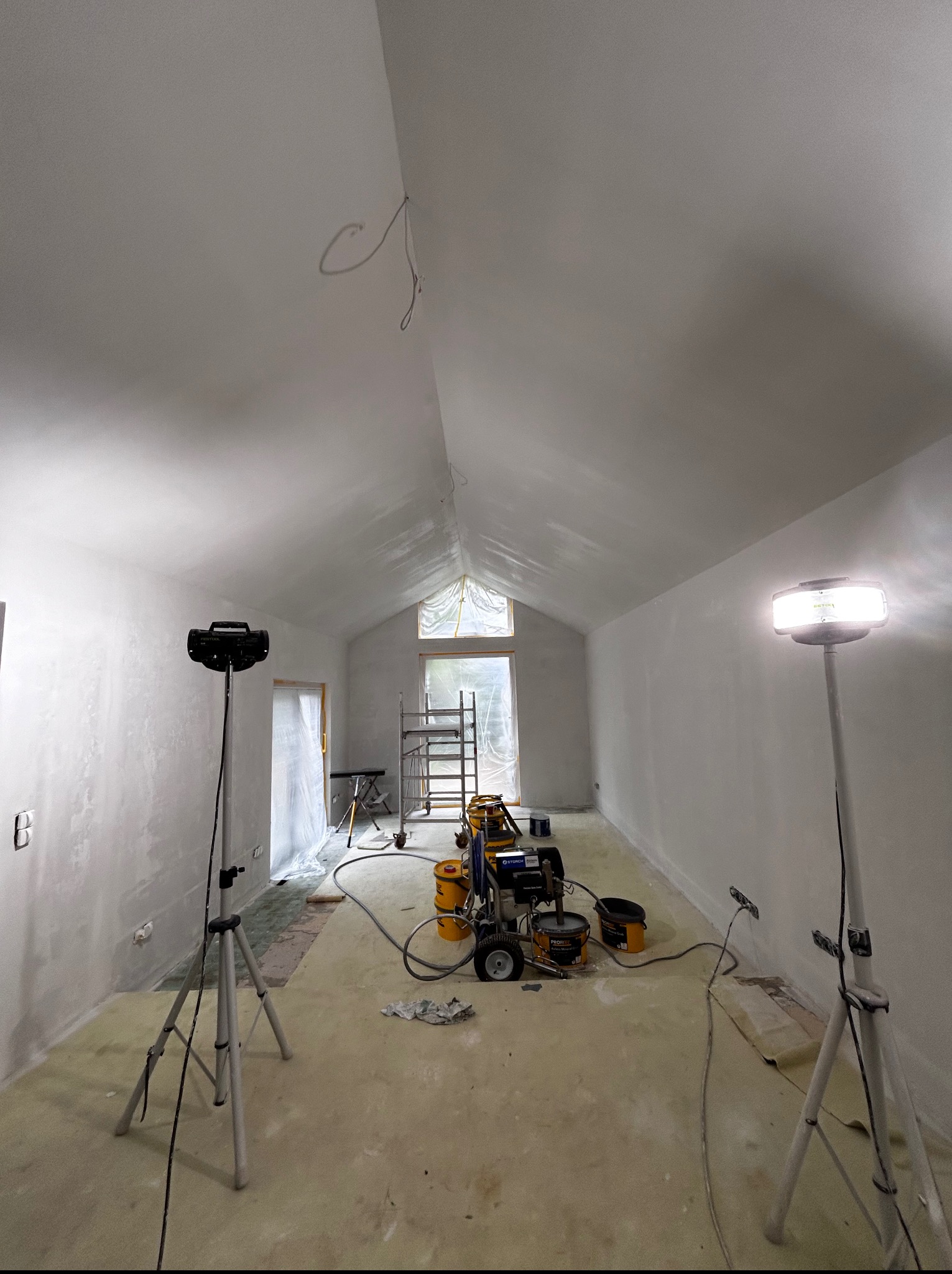 Wnętrze w trakcie malowania natryskowego na biało. Widoczny agregat malarski, reflektory na trójnogach i zabezpieczenia okna. Ściany i sufit pokryte świeżą farbą.