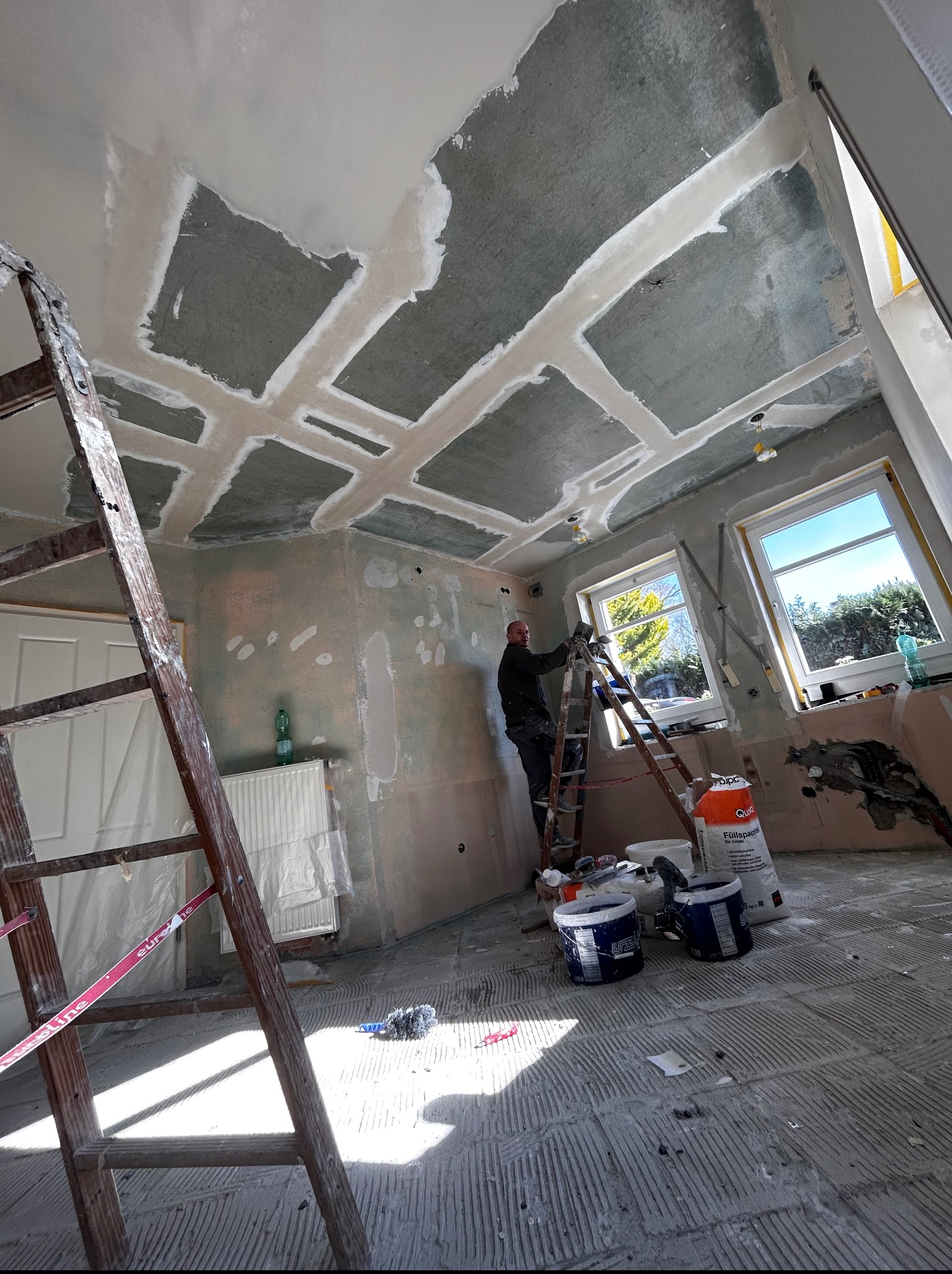 Prace szpachlarskie sufitu z widocznymi łączeniami płyt gipsowych, wykonywane przez mężczyznę na drabinie. W tle okna i widoczne narzędzia pracy.