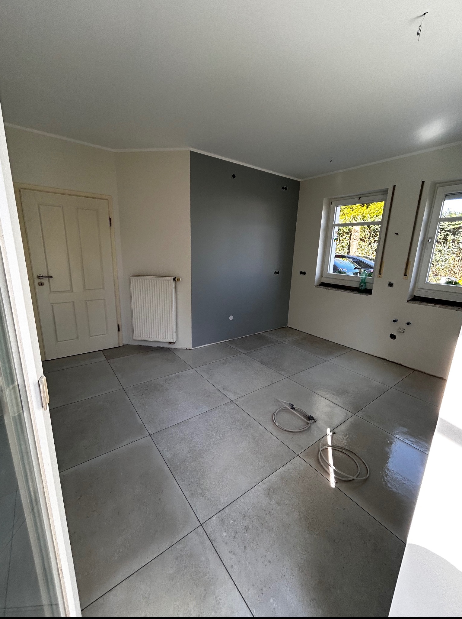 Wnętrze pokoju z szarą ścianą, grzejnikiem, oknami i szarymi płytkami podłogowymi. Widoczne przygotowania do dalszych prac wykończeniowych.