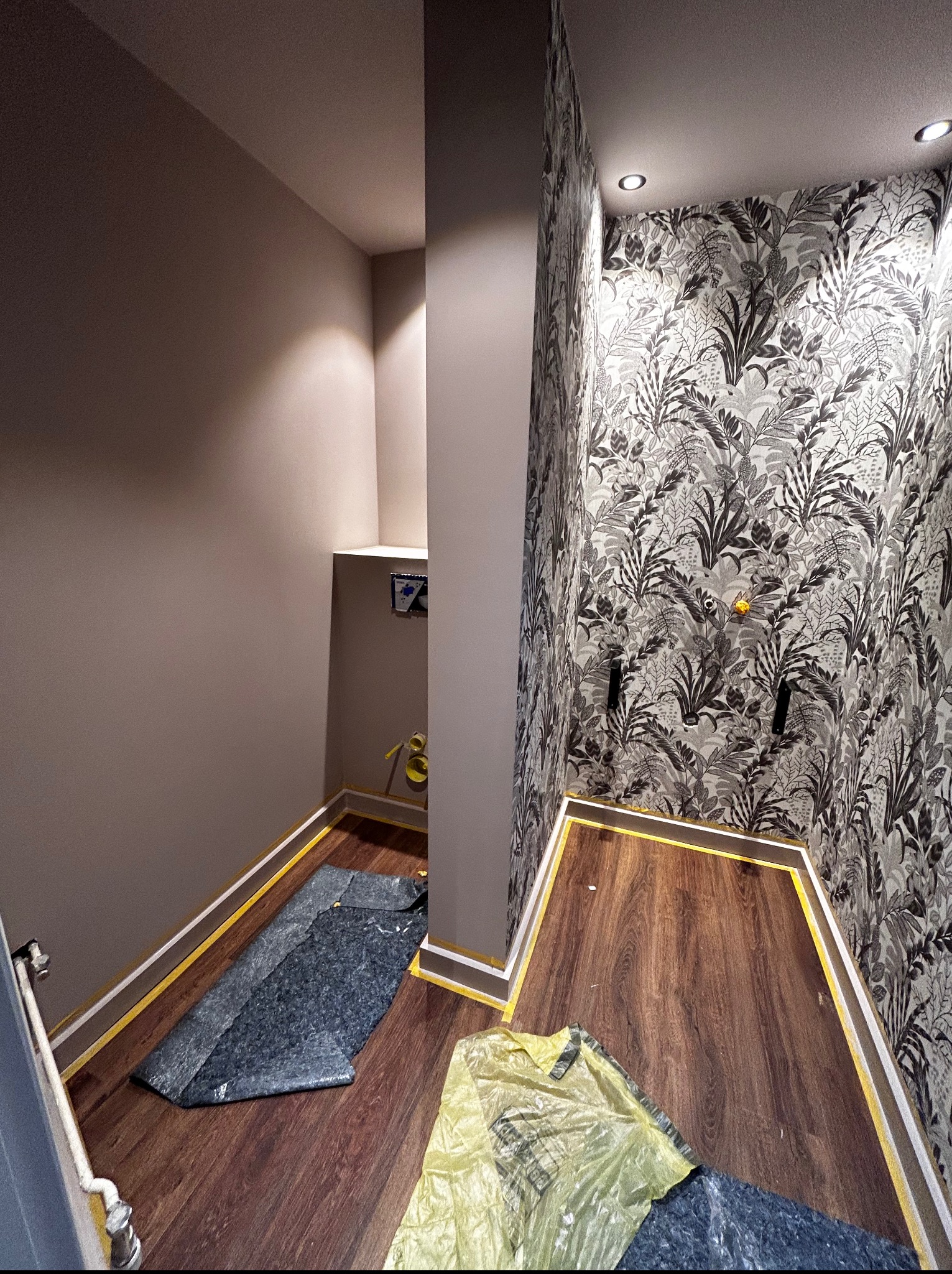 Wykończone wnętrze małej łazienki z drewnopodobną podłogą, tapetą w roślinny wzór i pomalowanymi na szaro ścianami. Zabezpieczona podłoga folią i taśmą malarską.