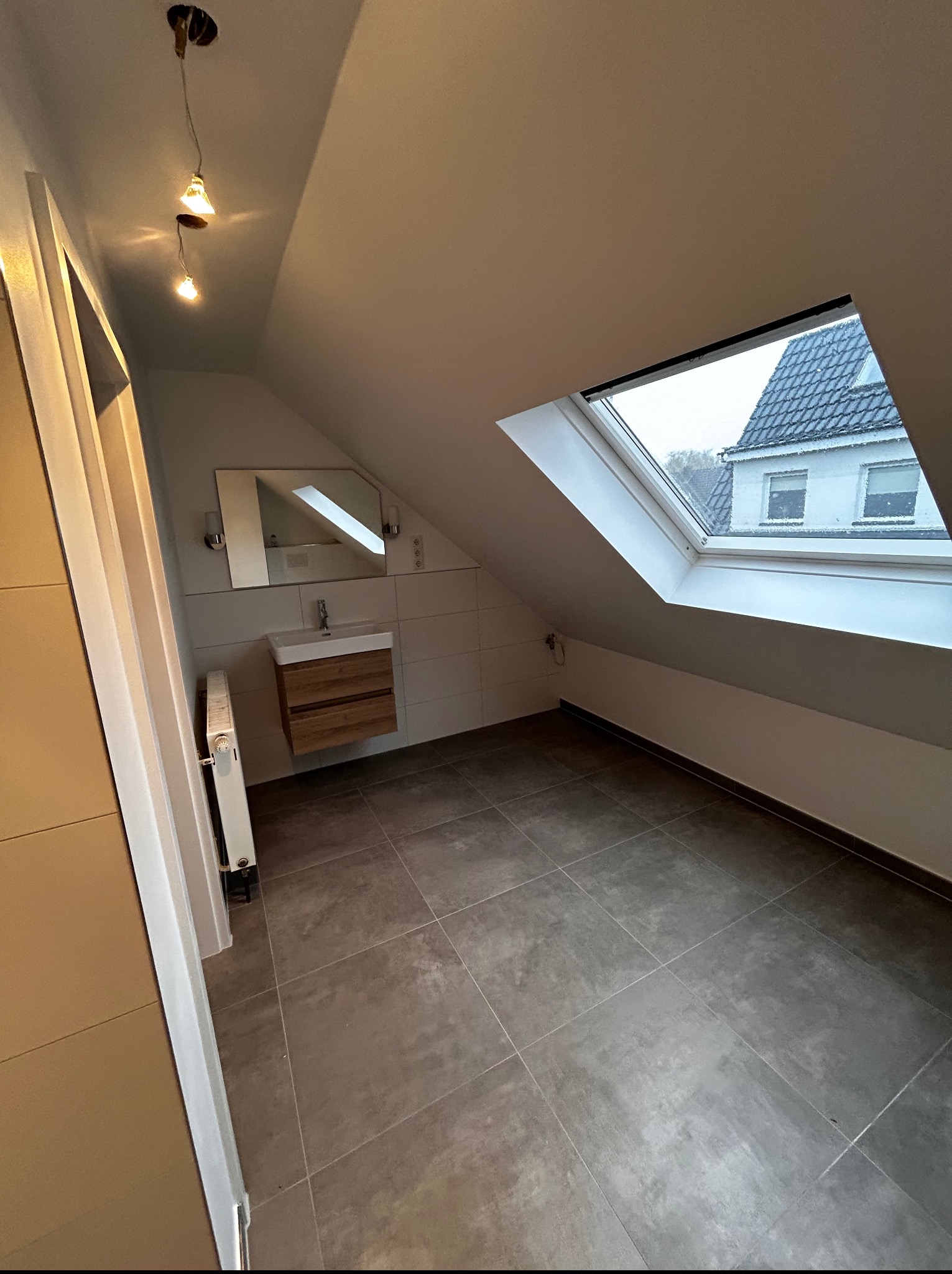 Nowoczesna łazienka na poddaszu z oknem dachowym, szarymi płytkami, drewnianą szafką pod umywalką i minimalistycznym oświetleniem. Widok na budynek przez okno.