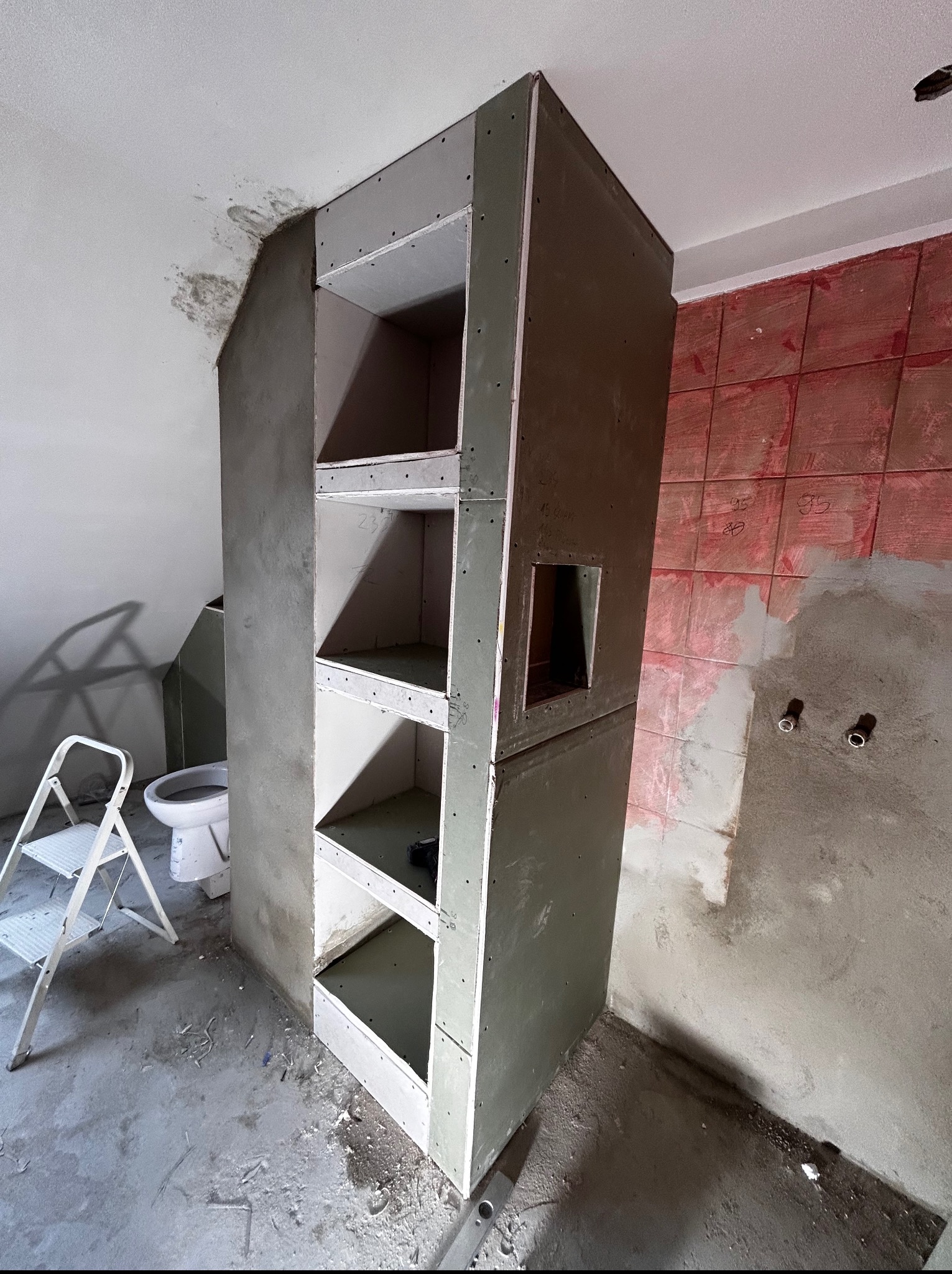 Wnętrze łazienki w trakcie remontu: konstrukcja z płyt gipsowo-kartonowych z półkami, drabina, surowe ściany i podłoga. Widoczne przygotowania do dalszych prac wykończeniowych.
