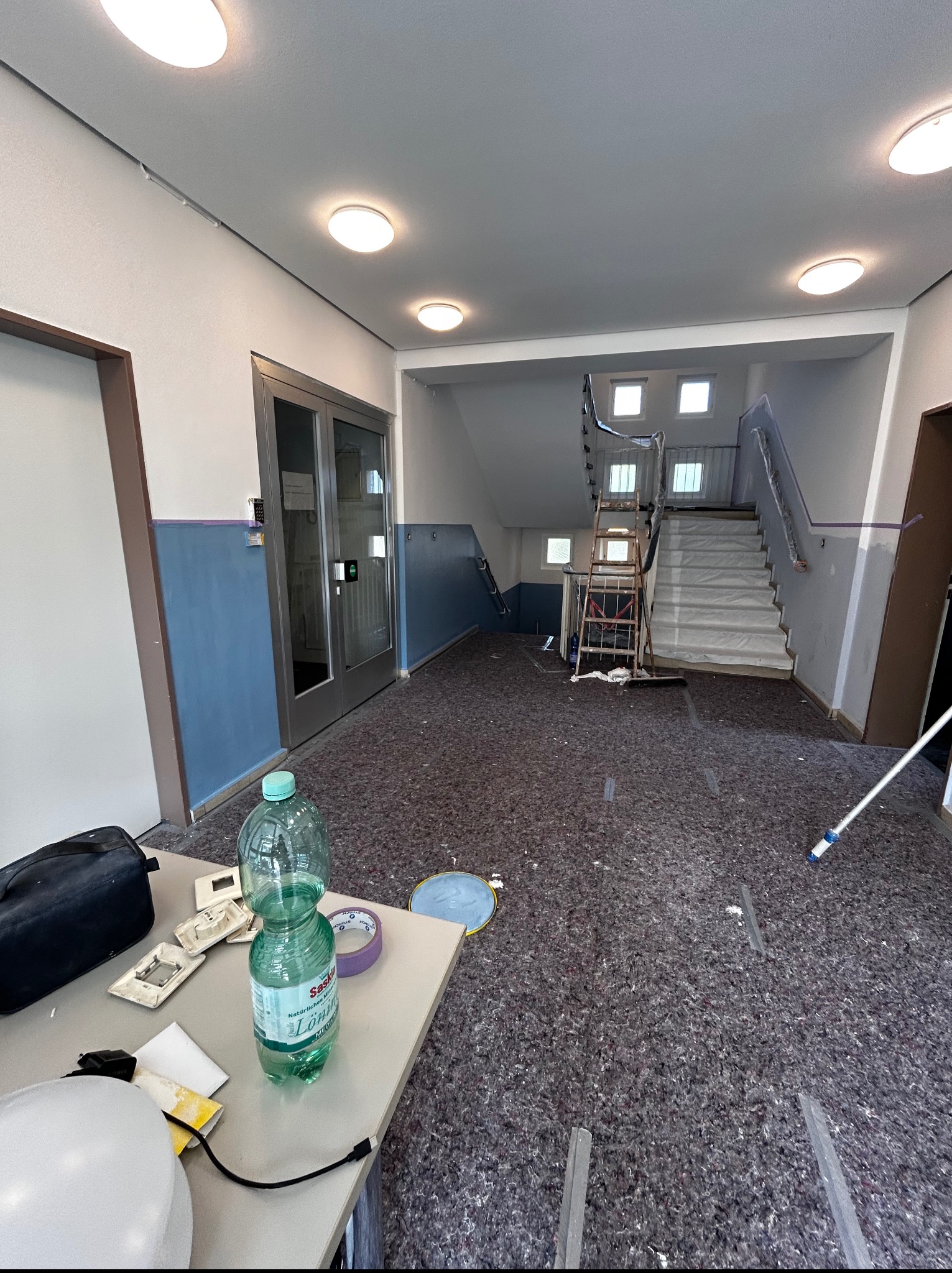 Korytarz w trakcie prac wykończeniowych: zabezpieczona podłoga, schody osłonięte folią, widoczna drabina i narzędzia malarskie. Przygotowania do malowania ścian.