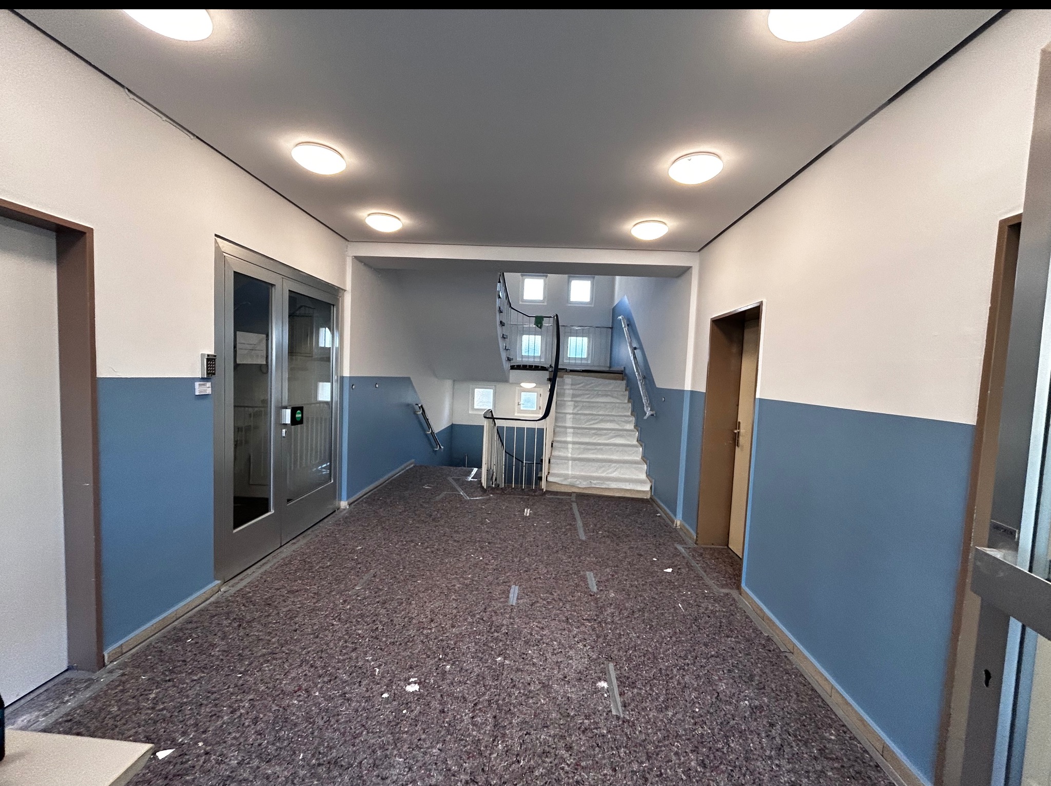 Korytarz w budynku po remoncie. Widoczne odmalowane ściany w kolorze biało-niebieskim, nowe oświetlenie sufitowe i zabezpieczone schody.