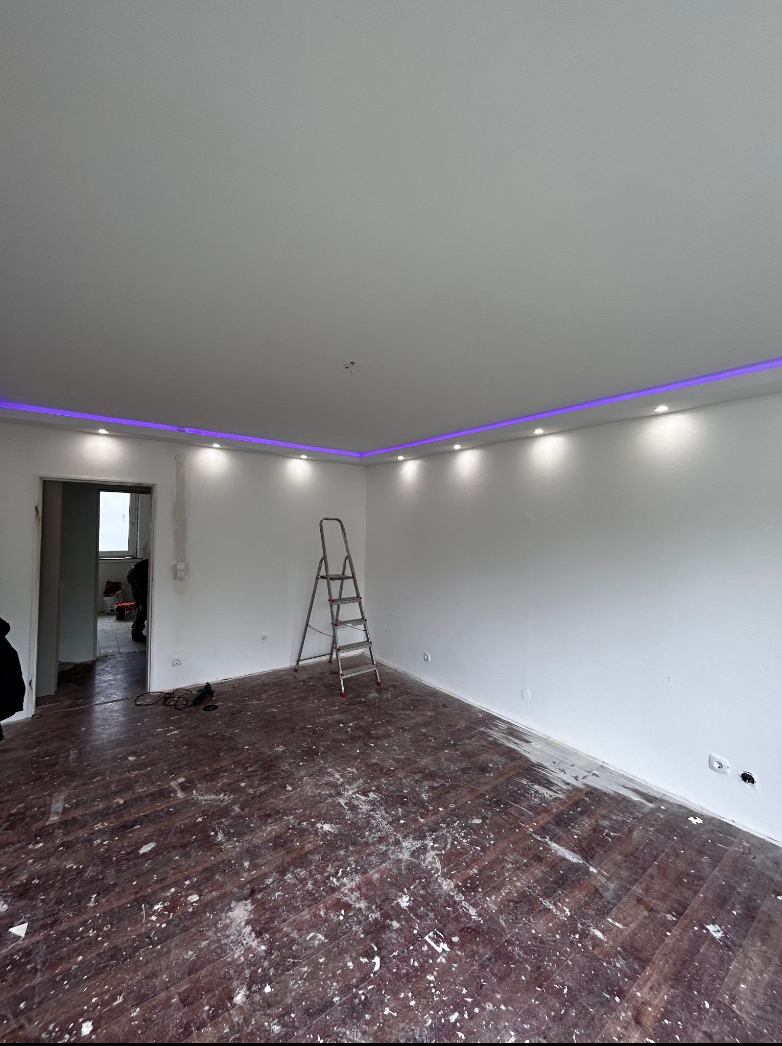 Białe wnętrze z zamontowanym oświetleniem LED przy suficie, drabina w rogu pokoju, ślady farby na podłodze, otwarte drzwi do kolejnego pomieszczenia.