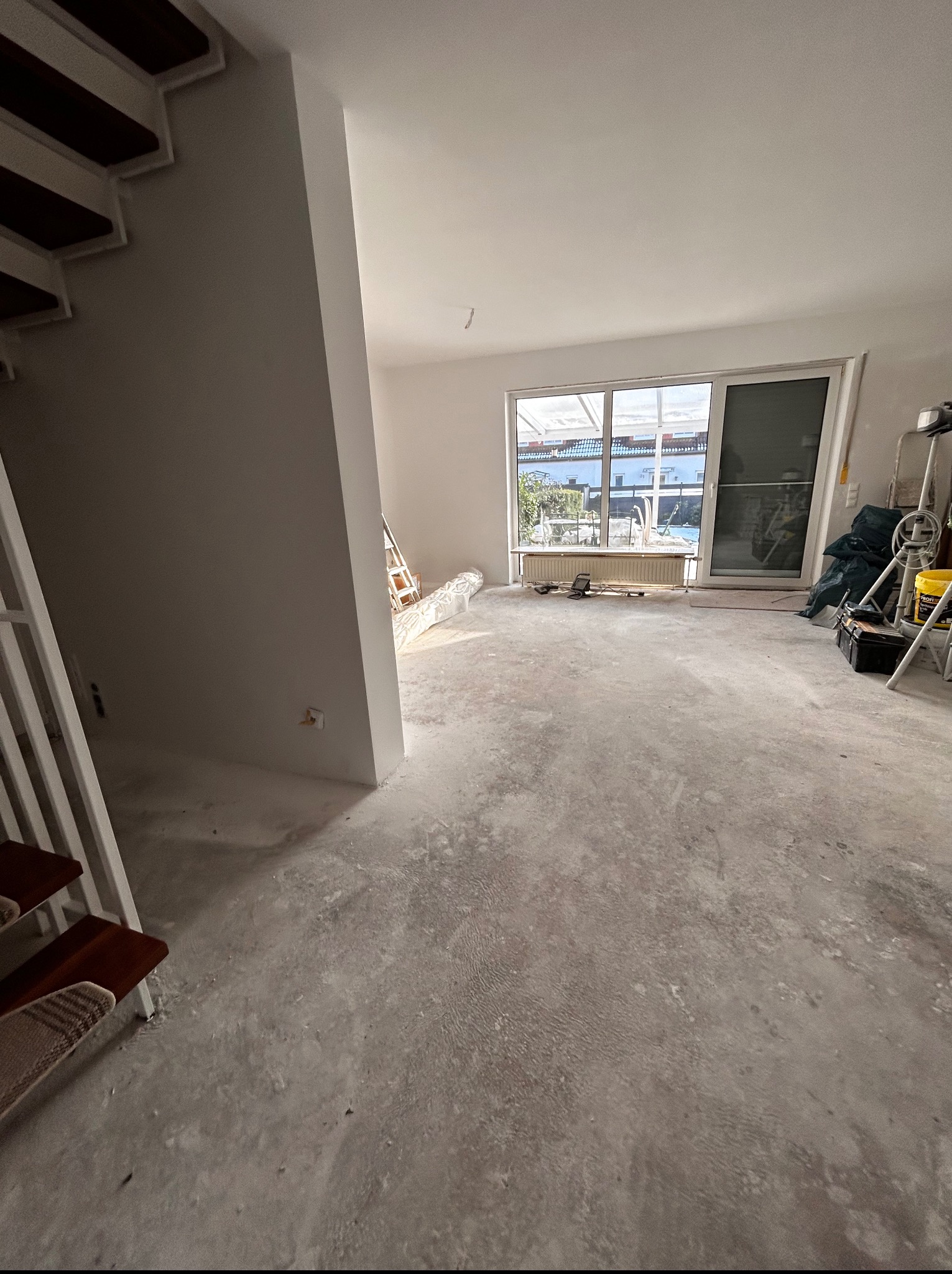Wnętrze w trakcie prac wykończeniowych: surowy beton na podłodze, fragment schodów i okno z widokiem na zewnątrz. Widoczne narzędzia i materiały budowlane.