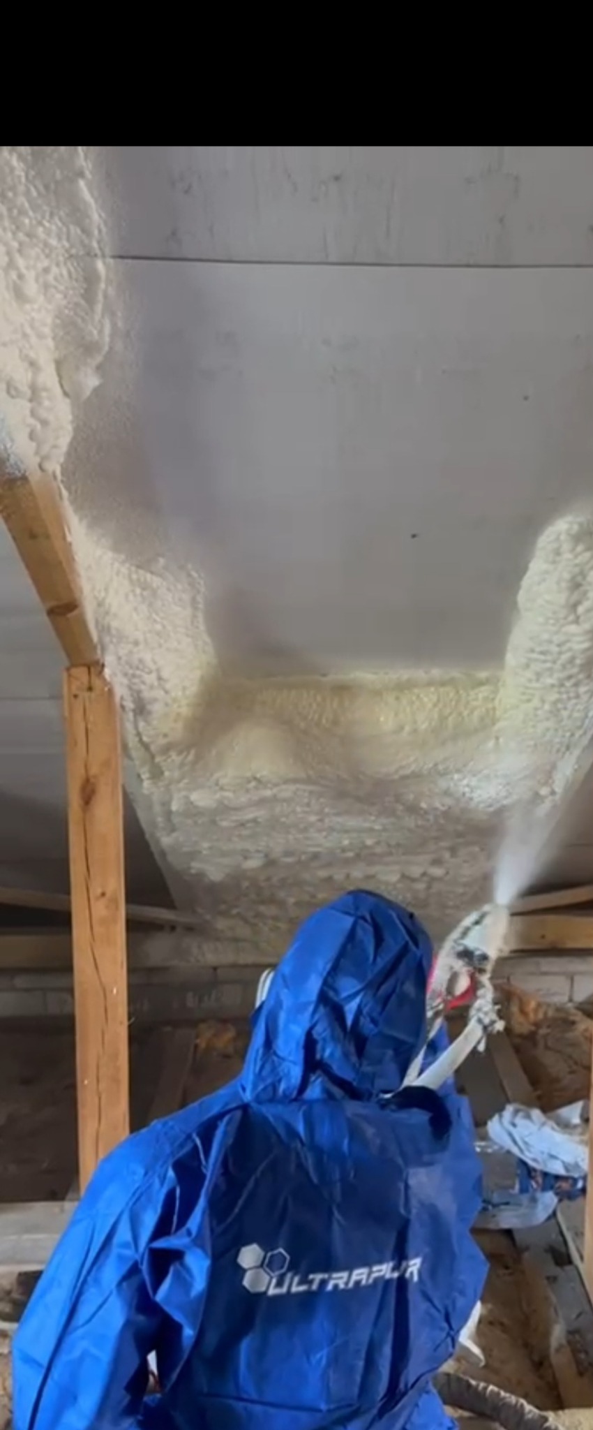 Aplikacja pianki PUR na poddaszu przez pracownika w niebieskim kombinezonie ochronnym, widoczne drewniane elementy konstrukcji dachu.