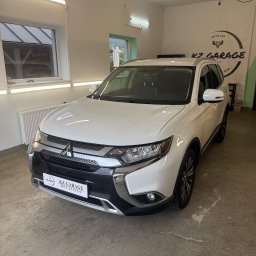 KZ Garage Krystian Zbozień - Biały Mitsubishi Outlander po detailingu w garażu KZ Garage, widok z przodu, logo firmy na tablicy rejestracyjnej, plakat reklamowy w tle.