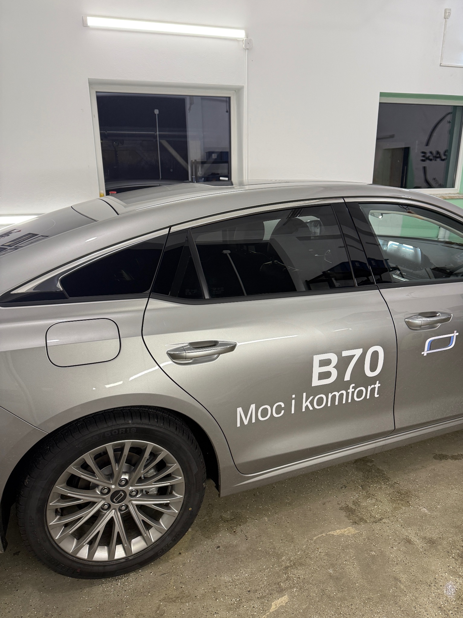 Srebrny sedan B70 z przyciemnionymi szybami, zaparkowany w jasnym garażu. Widoczne odbicie w oknach. Napis 'Moc i komfort' na karoserii.
