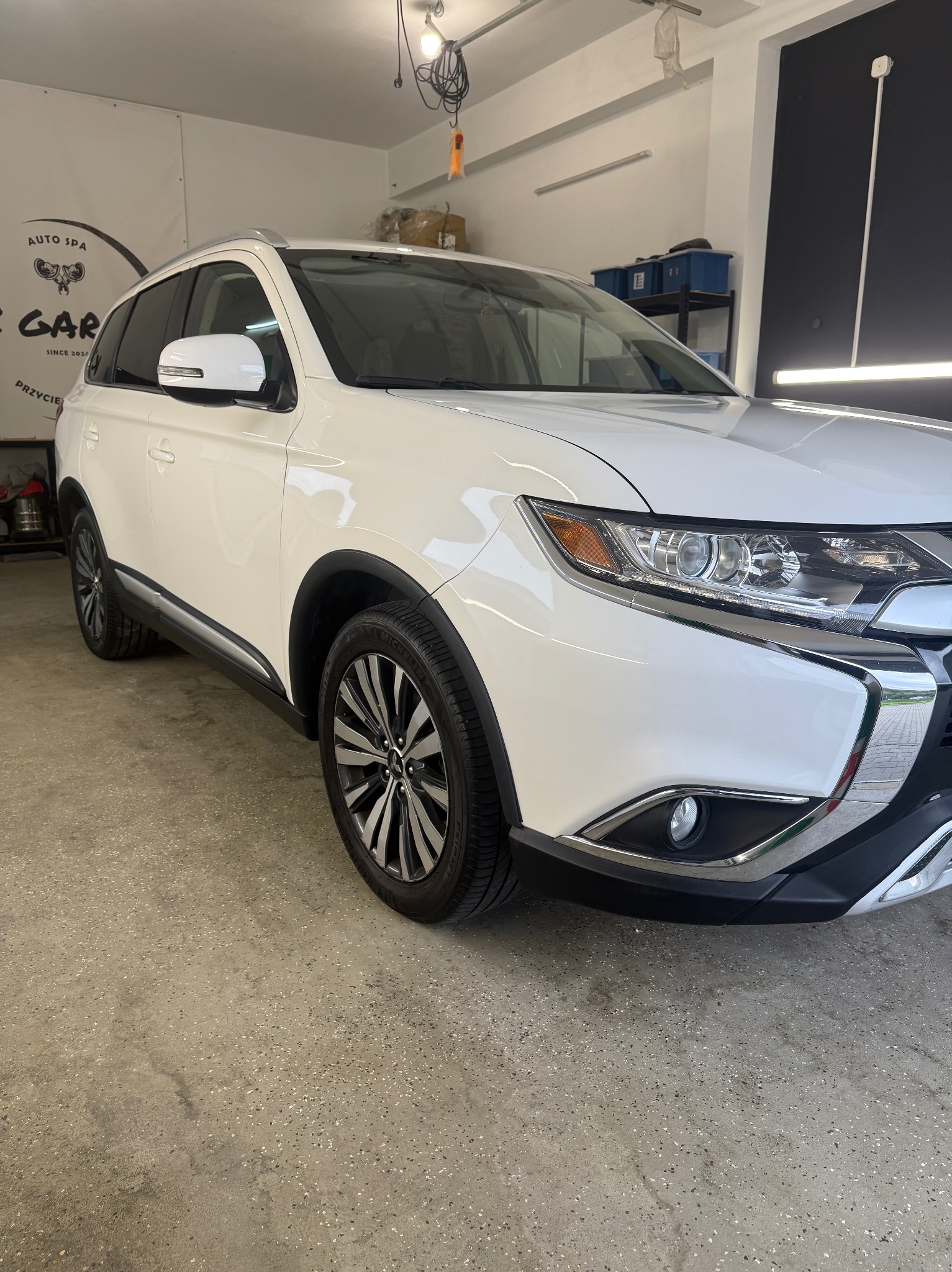 Biały SUV, Mitsubishi Outlander, po detailingu w garażu. Widoczne odblaski na lakierze, czyste felgi i opony. Auto ustawione pod kątem, z widocznym przodem i bokiem.