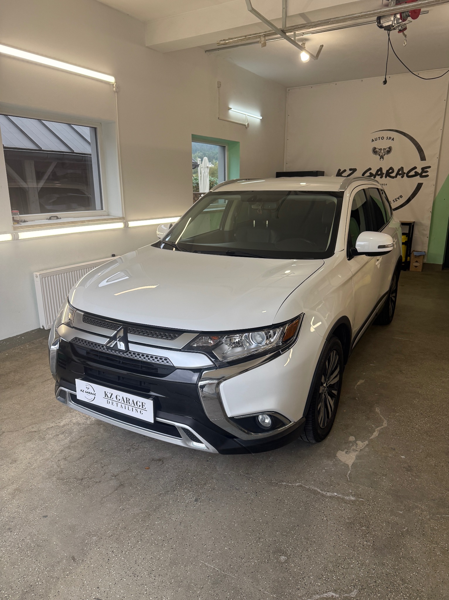 Biały Mitsubishi Outlander po detailingu w garażu KZ Garage, widok z przodu, logo firmy na tablicy rejestracyjnej, plakat reklamowy w tle.