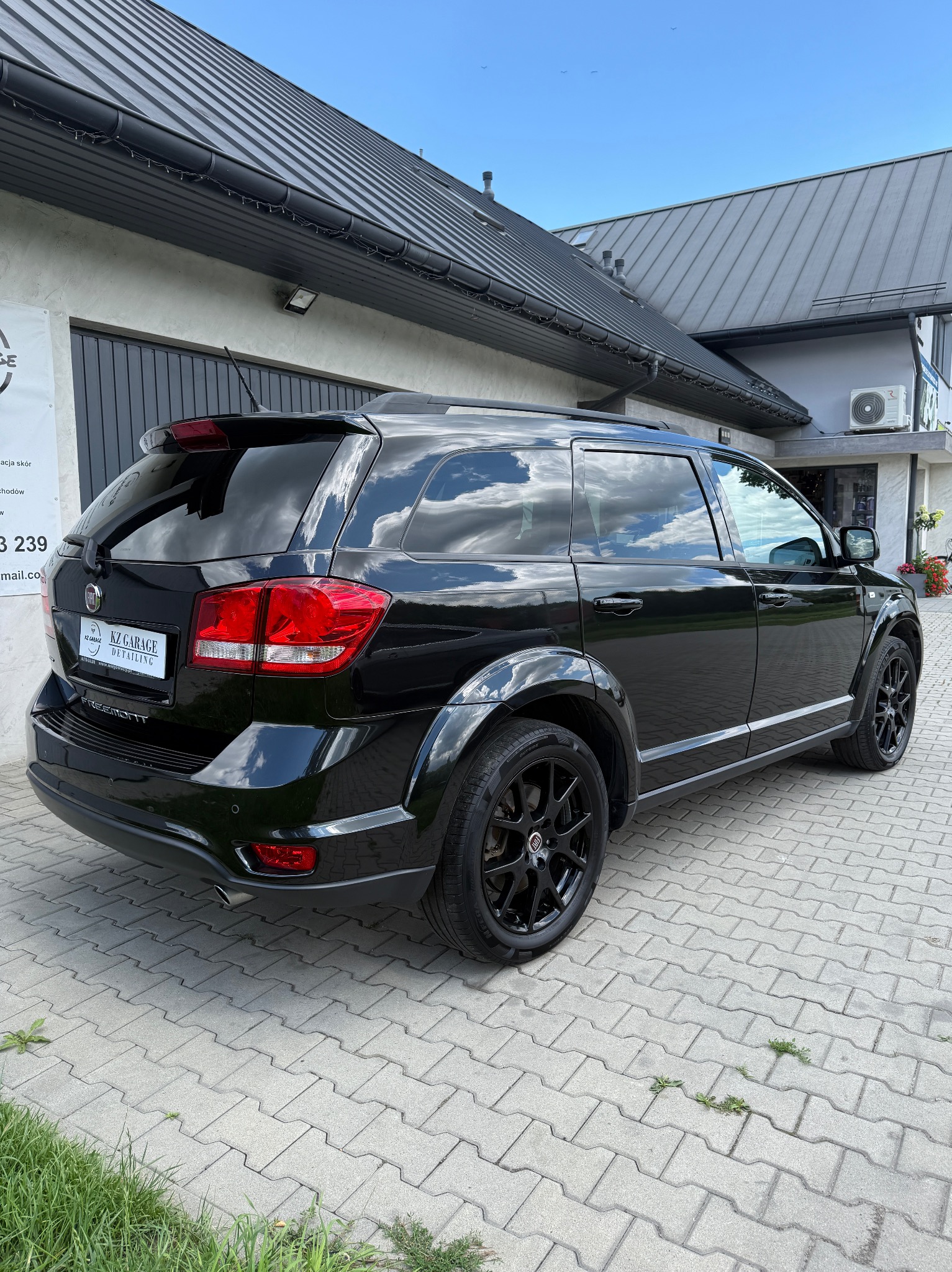 Czarny Fiat Freemont po detailingu, zaparkowany na kostce brukowej przed budynkiem. Widoczne odbicie chmur w lakierze i przyciemnione szyby.