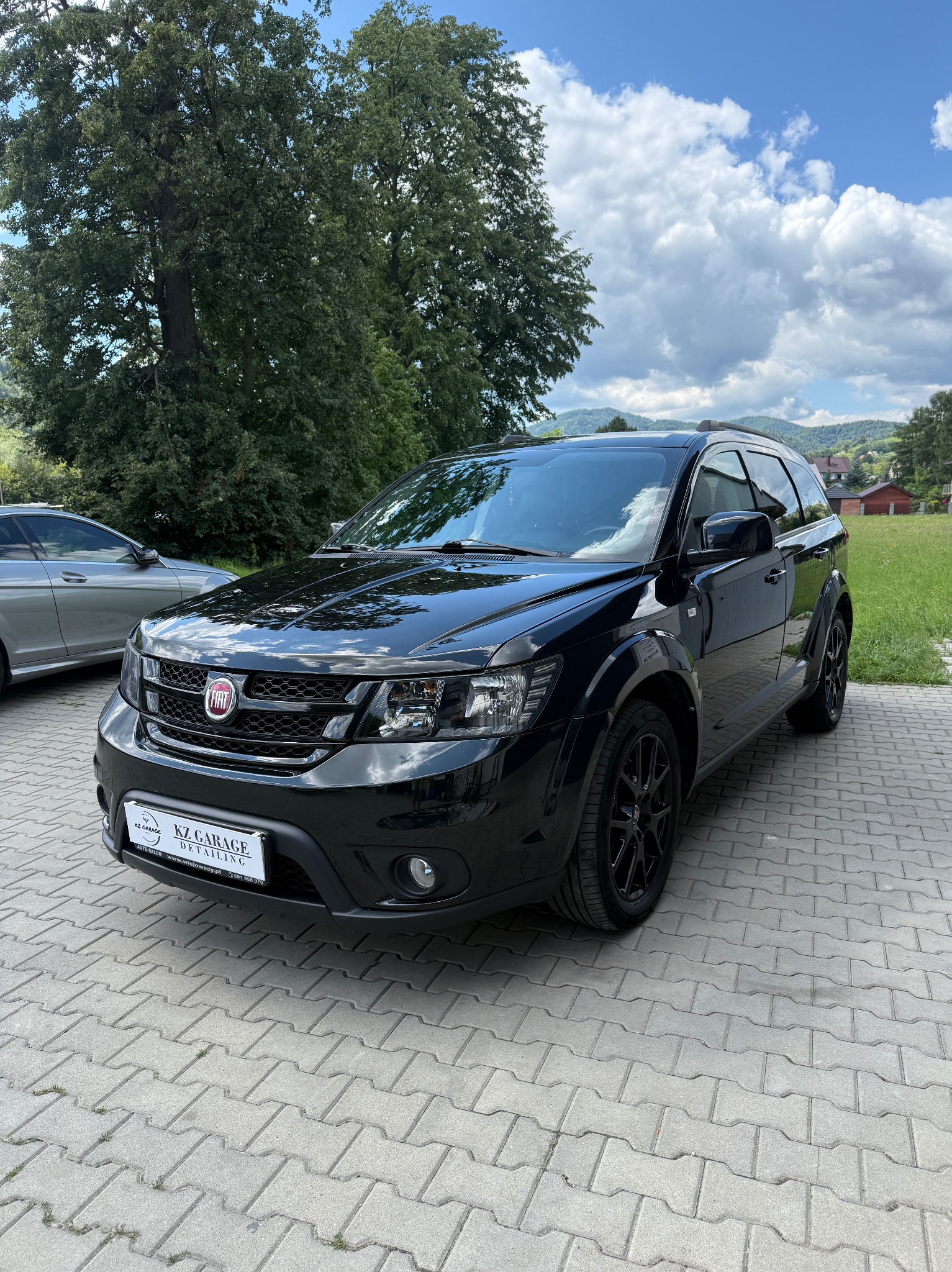 Błyszczący, czarny Fiat Journey po detailingu, zaparkowany na kostce brukowej. Widoczne logo KZ Garage Detailing. W tle zieleń drzew i budynki.