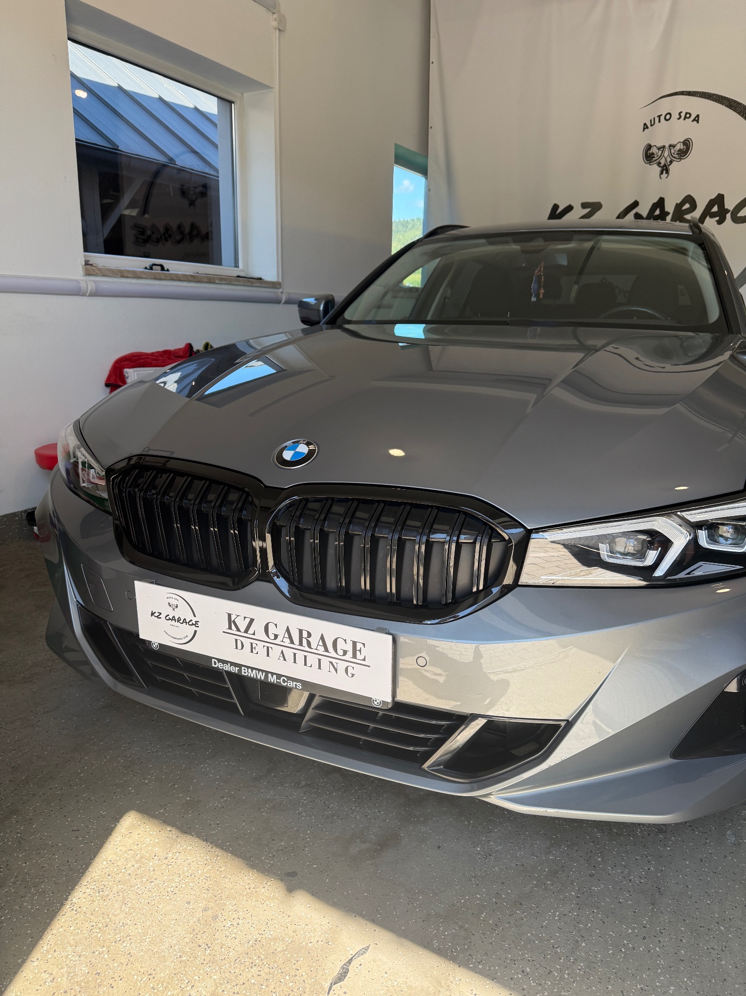Szary samochód BMW z czarną atrapą chłodnicy po detailingu w garażu KZ Garage, z logo firmy na tablicy rejestracyjnej i oświetleniem słonecznym na podłodze.