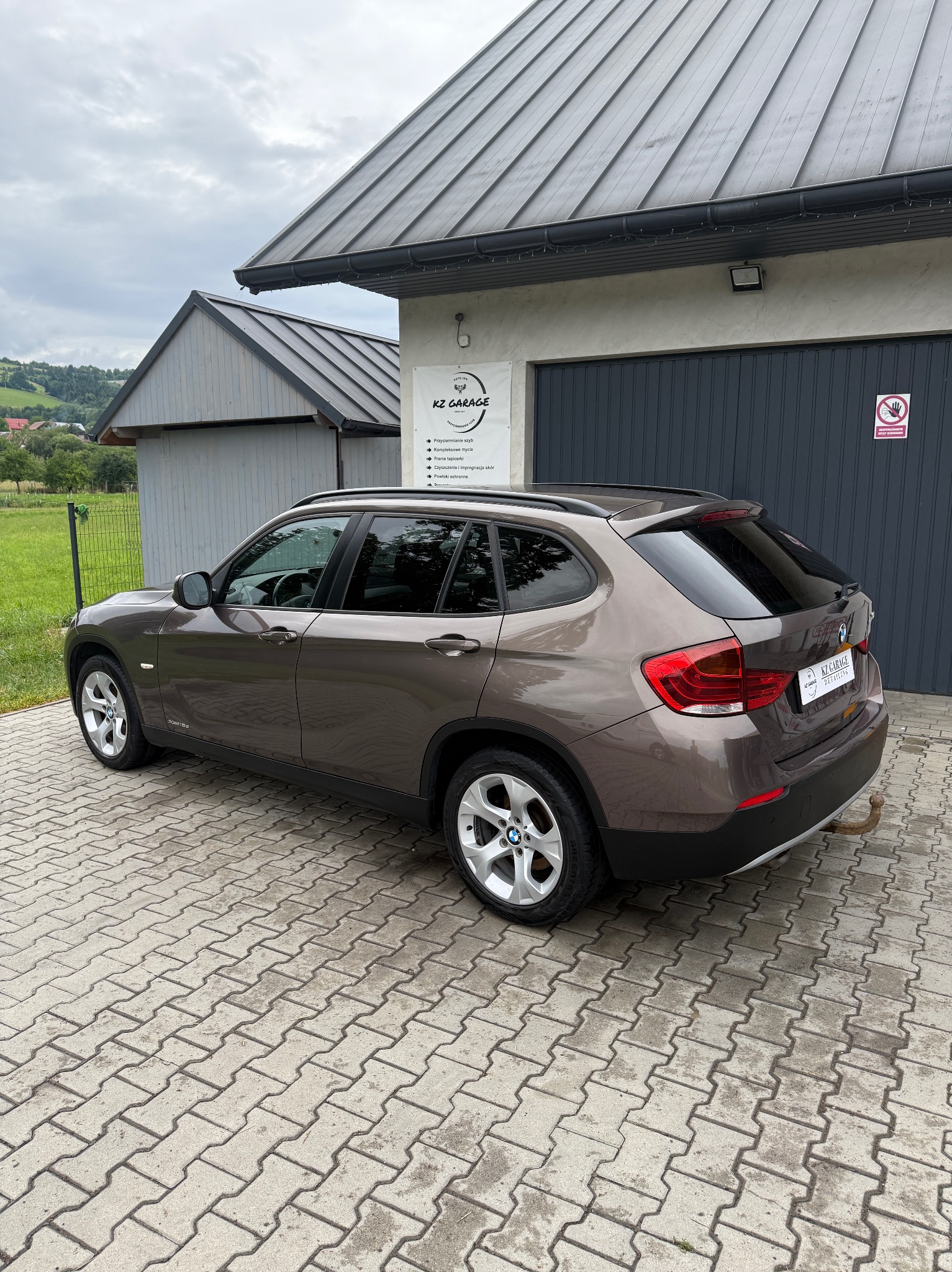 Brązowy SUV BMW X1 zaparkowany na kostce brukowej przed garażem z logo 'KZ Garage'. Widoczne przyciemnione szyby i hak holowniczy.