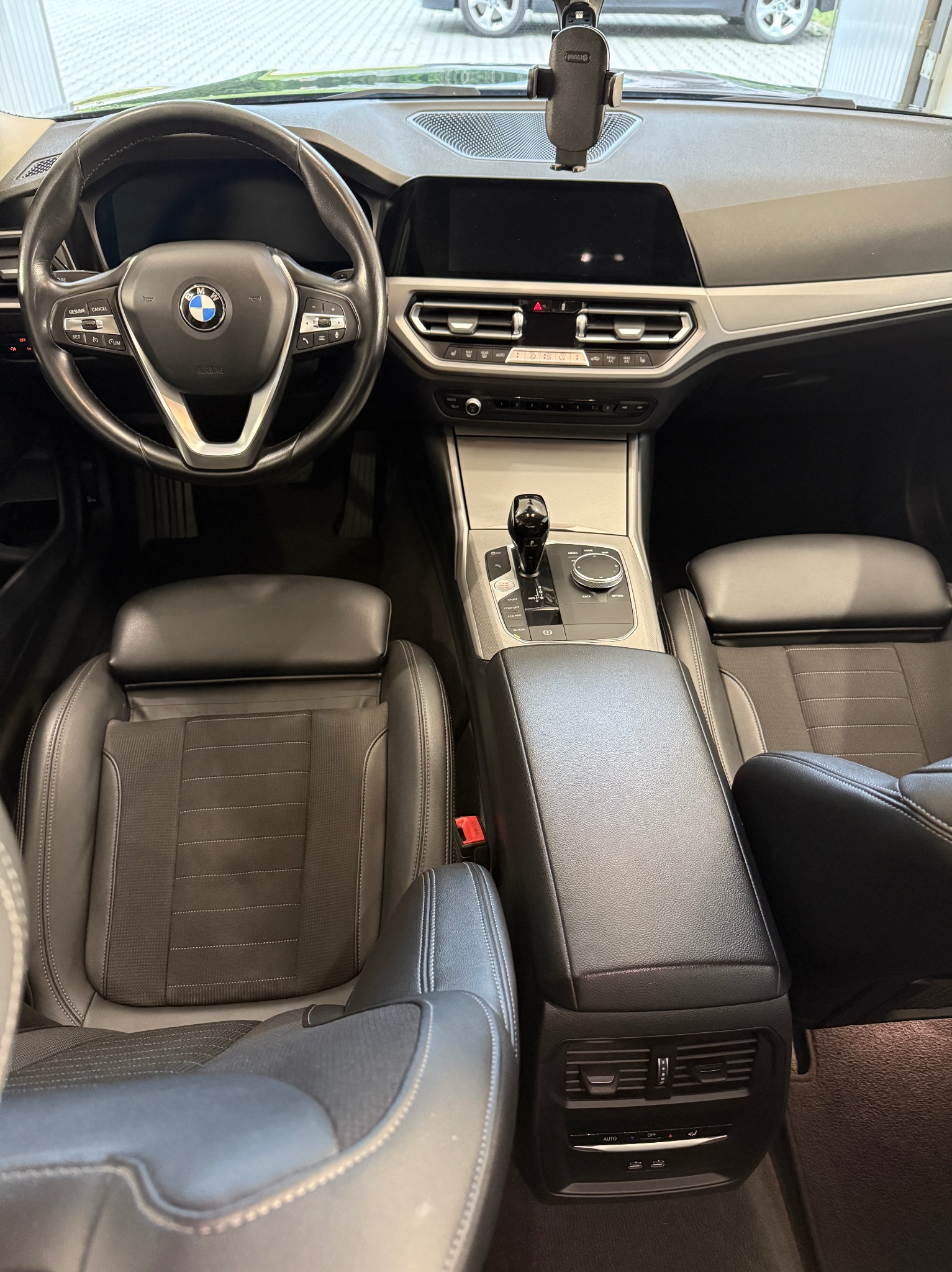 Wnętrze BMW serii 3 z czarną skórzaną tapicerką, widok na deskę rozdzielczą, kierownicę i fotele. W tle widoczny uchwyt na telefon i fragment szyby.
