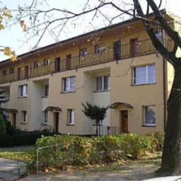 Rząd szeregowych domów jednorodzinnych z żółtą elewacją, czarnymi balkonami i małymi daszkami nad wejściami, otoczonych zielenią w jesiennym słońcu.