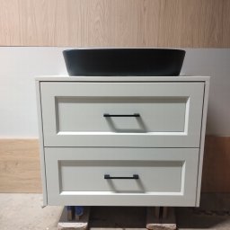 Qest meble - Biała szafka łazienkowa z dwiema szufladami, czarnymi uchwytami i umywalką na blacie, ustawiona na bloczkach w surowym wnętrzu.