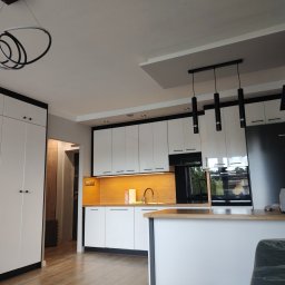 Qest meble - Nowoczesna, biała kuchnia z czarnymi akcentami: szafki, oświetlenie i duża zabudowa. Nietypowa lampa sufitowa. Widok na blat i okno.