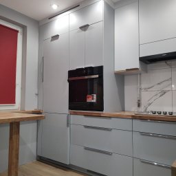 Qest meble - Jasnoszara zabudowa kuchenna z drewnianym blatem i czarnym piekarnikiem w nowoczesnym wnętrzu. Minimalistyczny design, widoczne detale wykończenia.