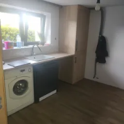 Funkcjonalna kuchnia z pralką pod blatem, szafka stojąca z jasnego drewna, okno z widokiem na zieleń. Minimalistyczny design, ciemna podłoga, surowa żarówka.