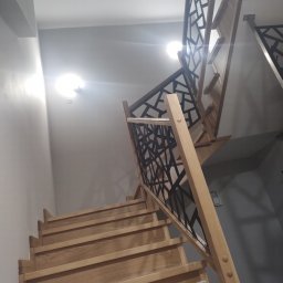 Qest meble - Drewniane schody z geometryczną, czarną balustradą, widok z dołu. Jasne drewno kontrastuje z ciemnym wzorem balustrady. Oświetlenie sufitowe.