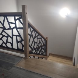 Qest meble - Drewniane schody z geometryczną, czarną balustradą. Widok z góry na pierwsze stopnie, łączące ciemną podłogę z jasnymi stopniami. Minimalistyczny design.