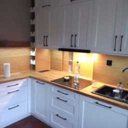 Biała kuchnia na wymiar z drewnianym blatem i czarnymi uchwytami. Widoczne szafki górne i dolne, płyta indukcyjna, zlew i fragment podłogi z brązowych płytek.