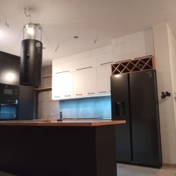 Qest meble - Nowoczesna kuchnia w trakcie montażu: czarne szafki, białe fronty, wyspa z drewnianym blatem, czarna lodówka i regał na wino. Widoczne niedokończone wykończenia.