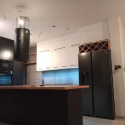 Nowoczesna kuchnia w trakcie montażu: czarne szafki, białe fronty, wyspa z drewnianym blatem, czarna lodówka i regał na wino. Widoczne niedokończone wykończenia.