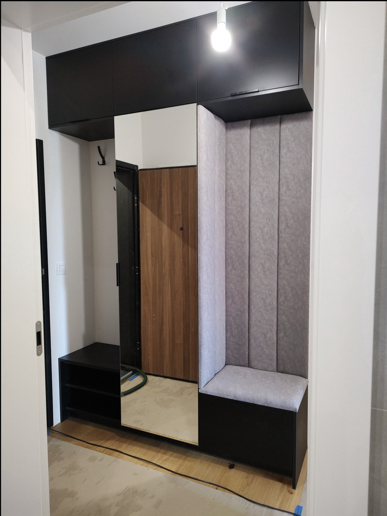 Funkcjonalna zabudowa przedpokoju z szafkami, lustrem i tapicerowanym siedziskiem w szaro-czarnej kolorystyce. Nowoczesny design, oświetlenie punktowe, widoczny fragment podłogi.
