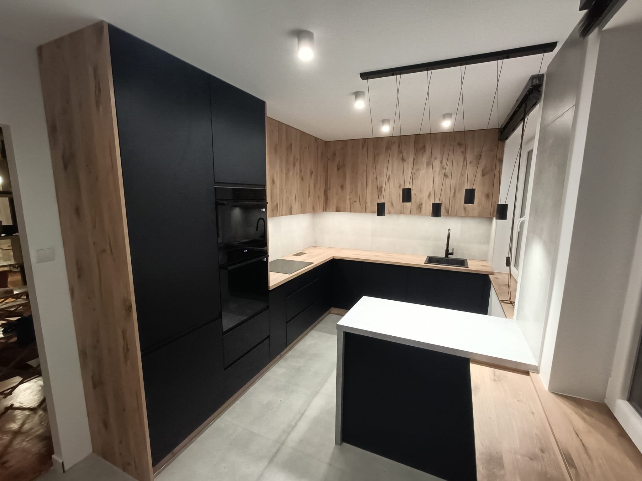 Nowoczesna kuchnia z czarnymi szafkami i drewnianymi akcentami, wyspą z białym blatem oraz oświetleniem punktowym i wiszącym. Minimalistyczny design.