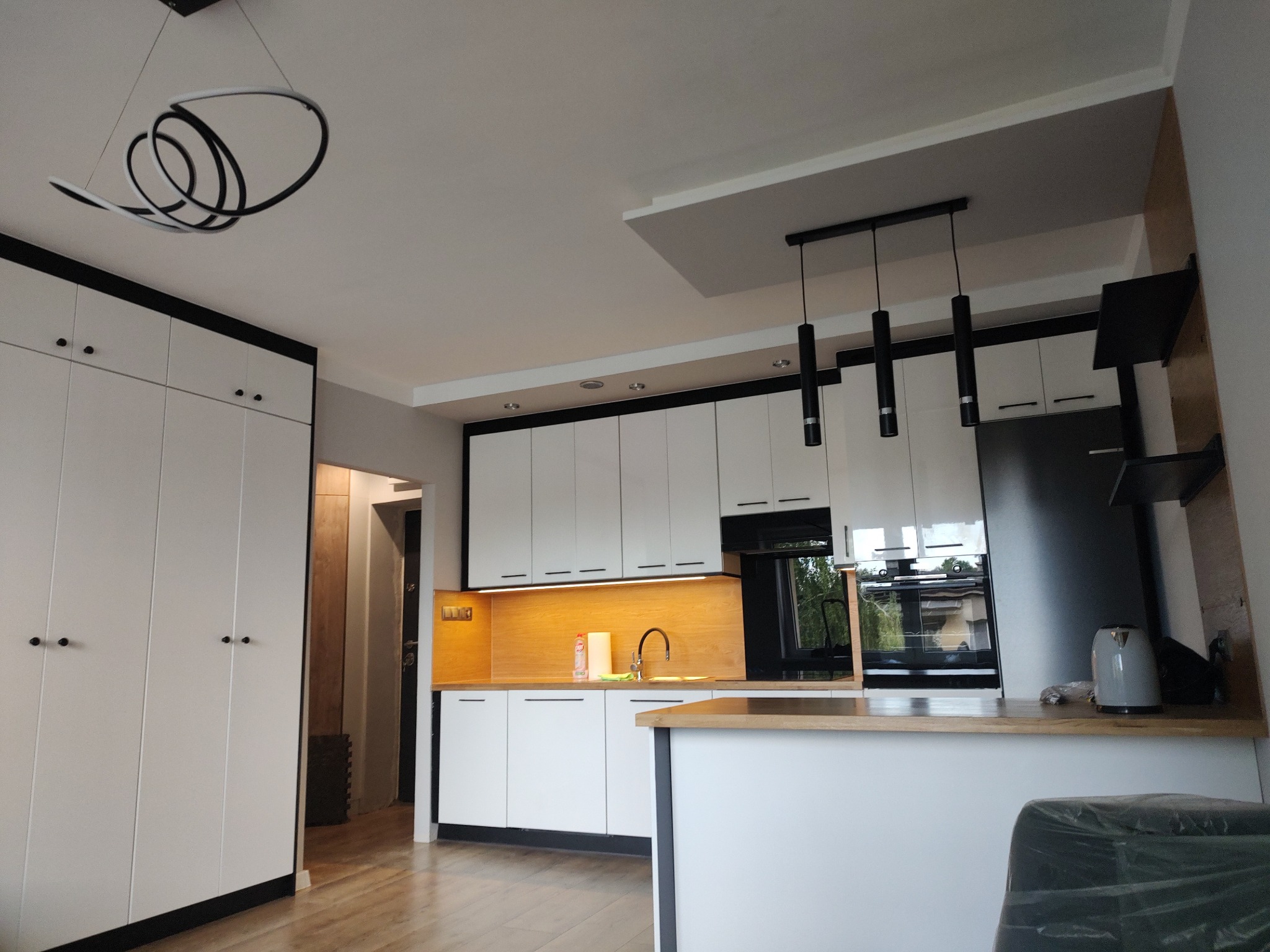 Nowoczesna, biała kuchnia z czarnymi akcentami: szafki, oświetlenie i duża zabudowa. Nietypowa lampa sufitowa. Widok na blat i okno.