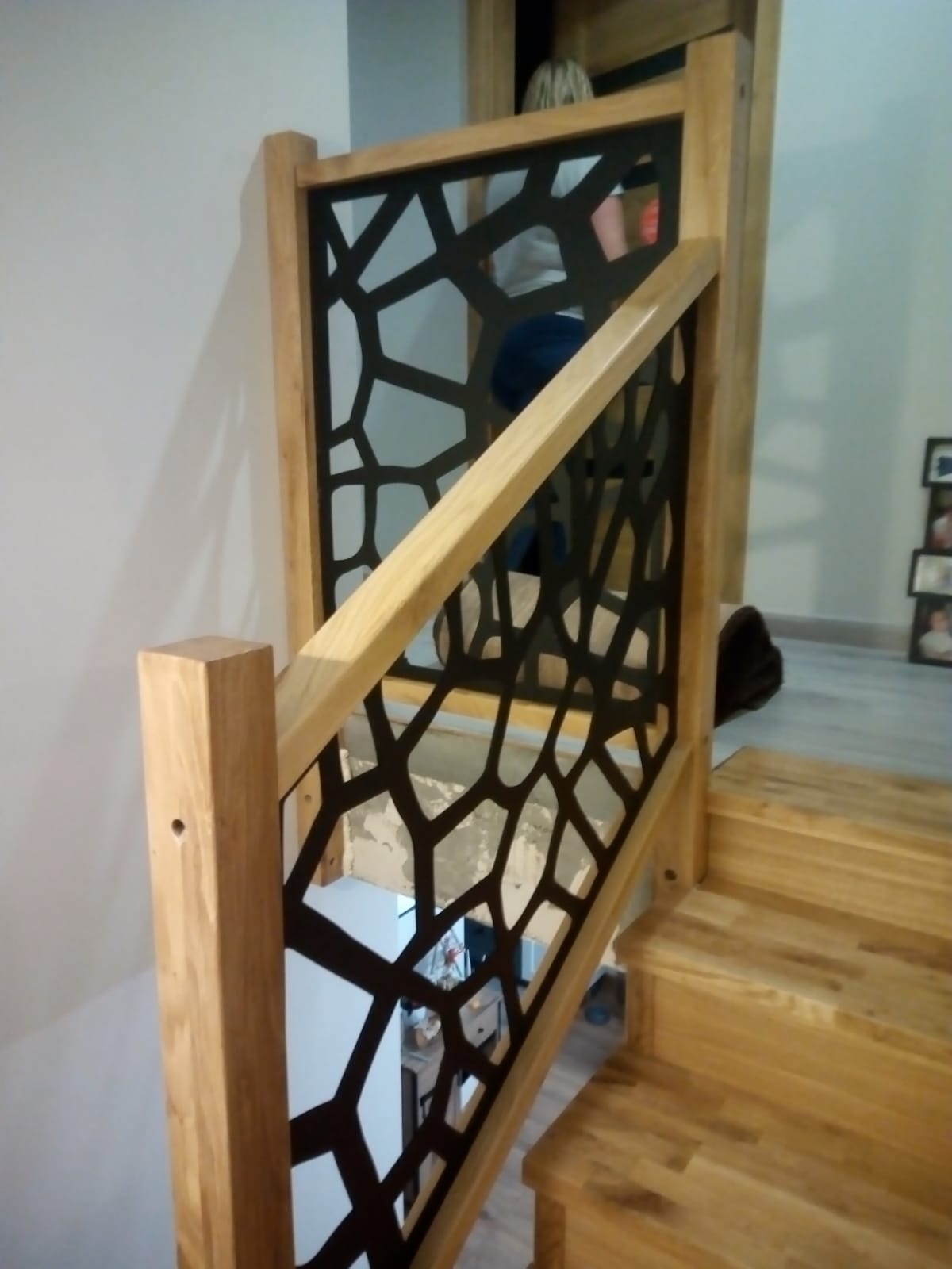 Drewniane schody z nowoczesną balustradą o geometrycznym wzorze. Jasne drewno i czarne elementy tworzą kontrast. Widok z góry na stopnie.