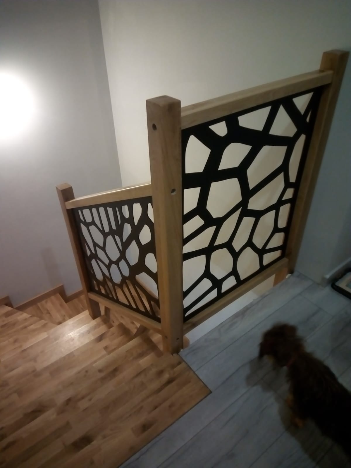 Drewniane schody z ozdobną balustradą o geometrycznym wzorze, widok z góry. Jasne drewno i czarne wypełnienie tworzą nowoczesny design.