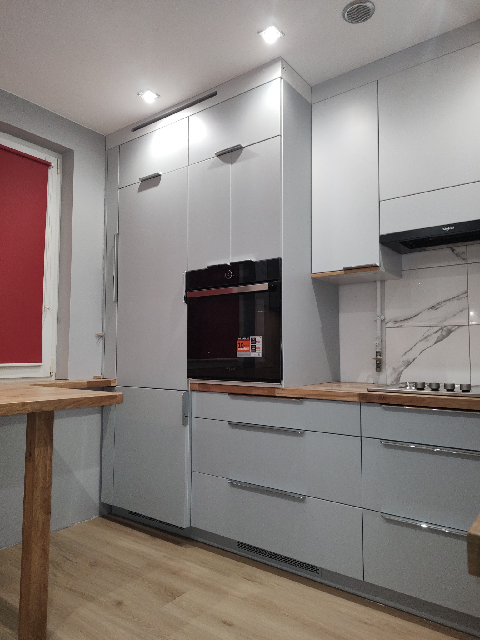 Jasnoszara zabudowa kuchenna z drewnianym blatem i czarnym piekarnikiem w nowoczesnym wnętrzu. Minimalistyczny design, widoczne detale wykończenia.