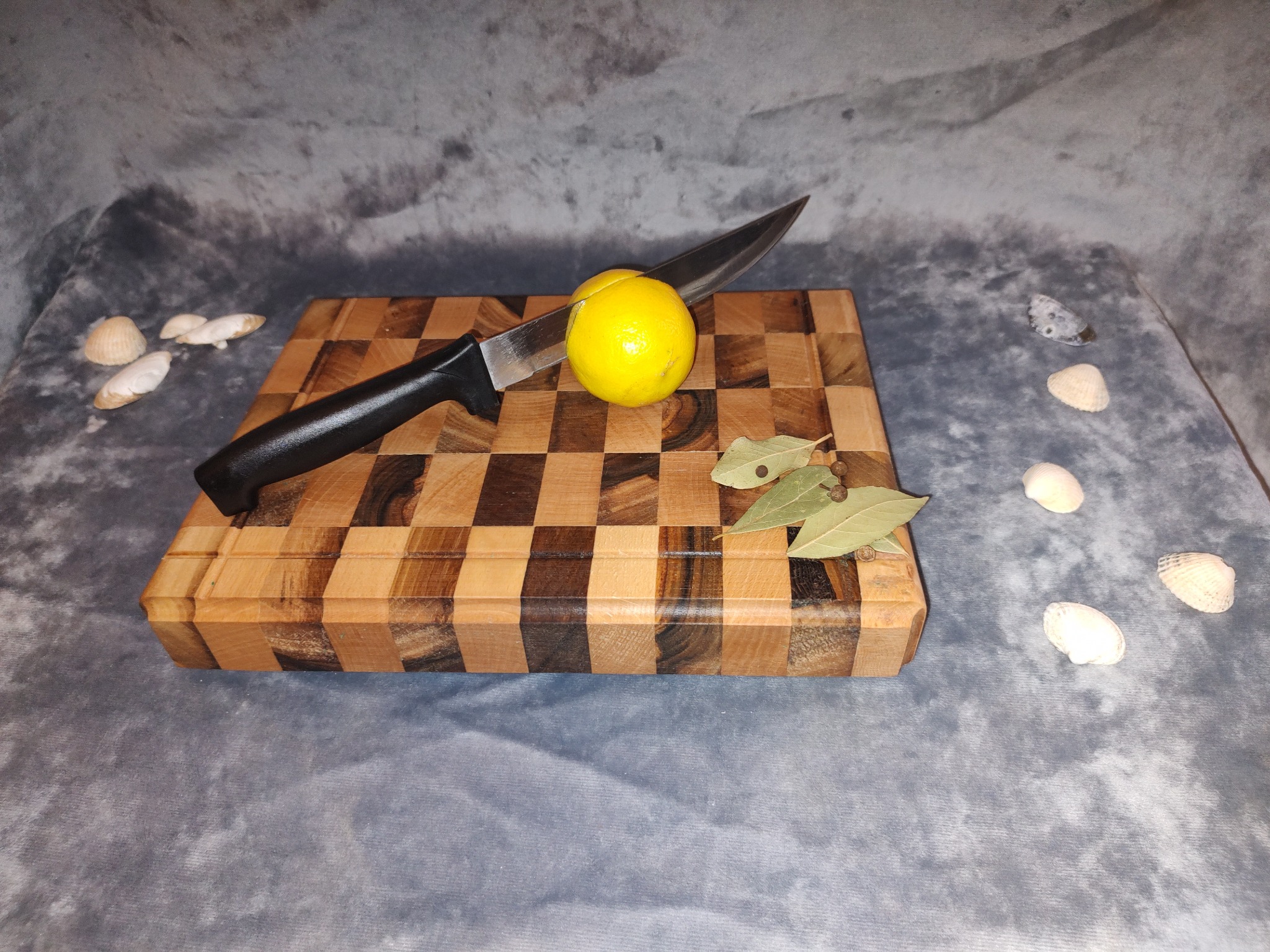 Deska do krojenia z szachownicą, cytryna przekrojona nożem, liście laurowe i muszelki na szarym tle.
