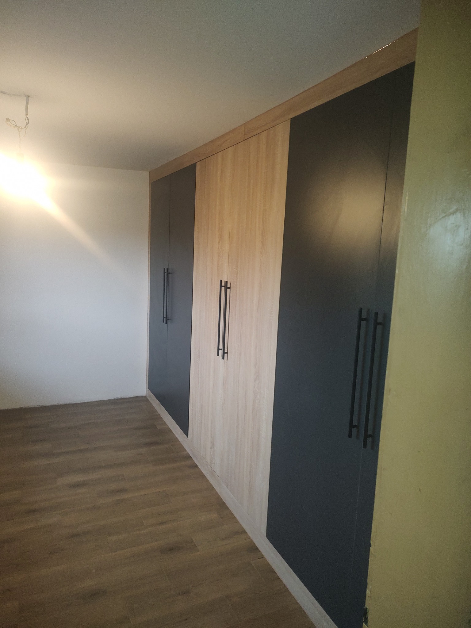 Długa szafa wnękowa z frontami w odcieniach drewna i grafitu, z długimi, czarnymi uchwytami, ustawiona wzdłuż ściany z drewnianą podłogą.