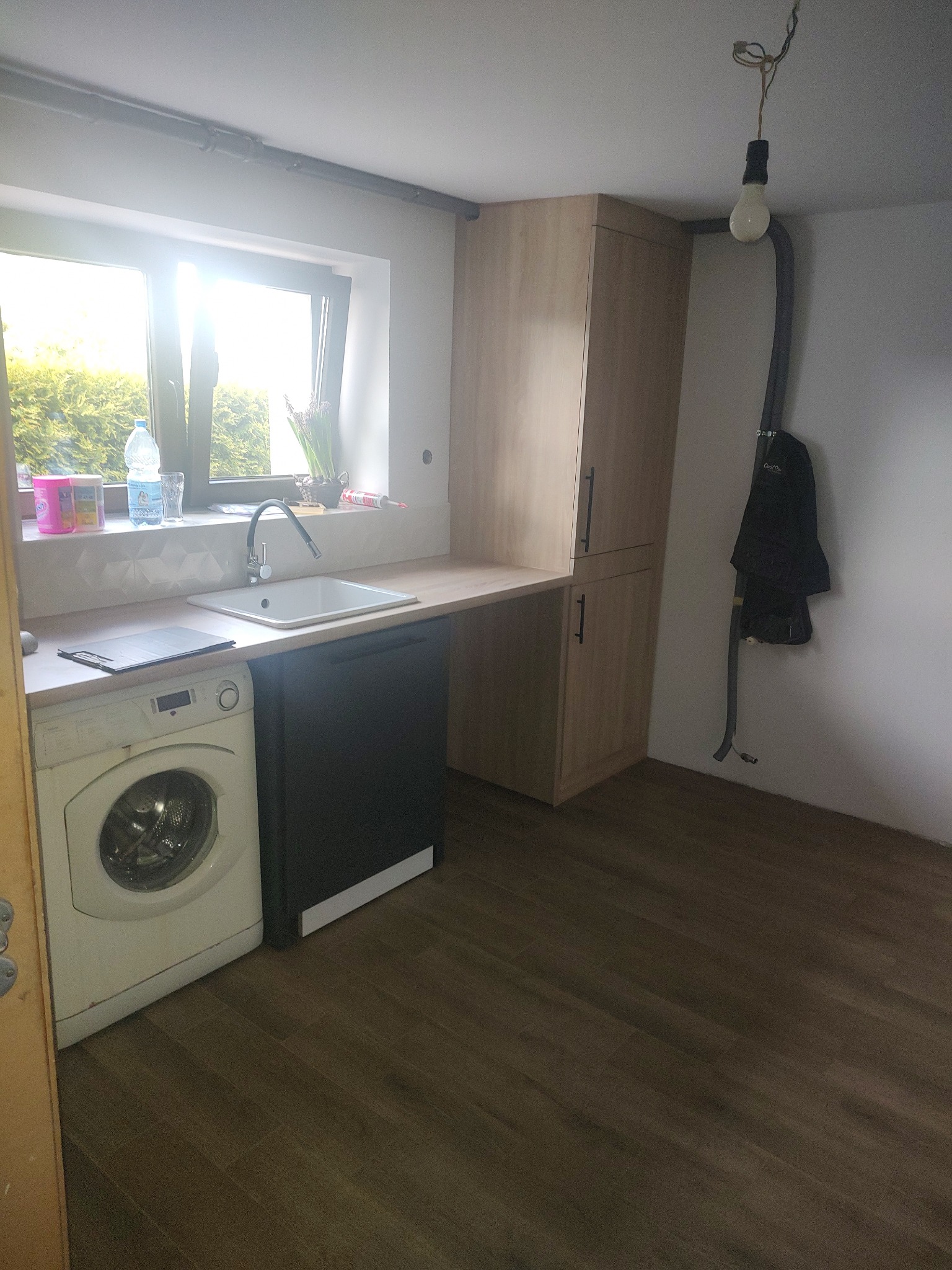 Funkcjonalna kuchnia z pralką pod blatem, szafka stojąca z jasnego drewna, okno z widokiem na zieleń. Minimalistyczny design, ciemna podłoga, surowa żarówka.