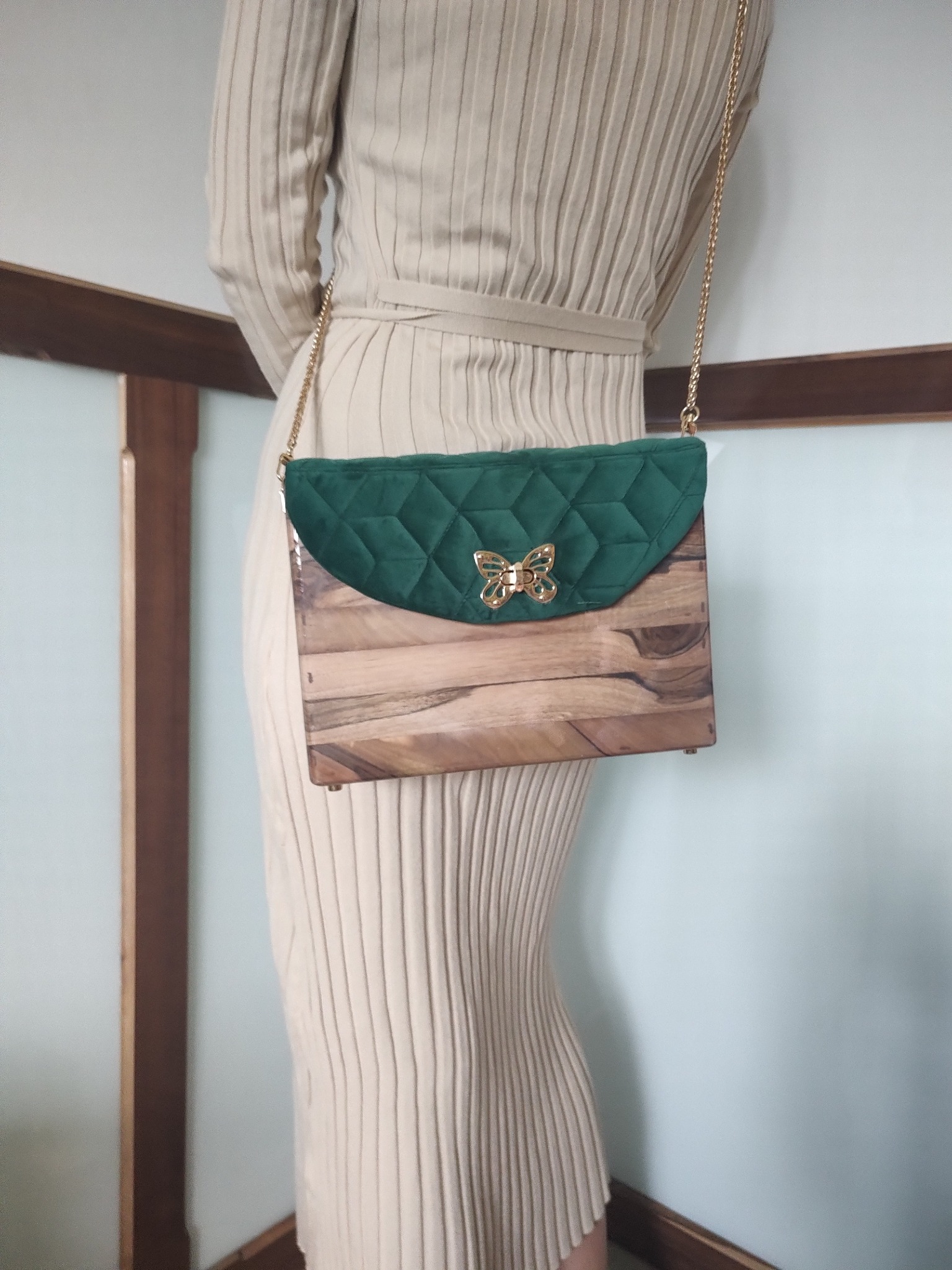 Kobieta prezentuje drewnianą torebkę z zielonym, pikowanym zapięciem i złotym motylem. Torebka zawieszona na złotym łańcuszku.