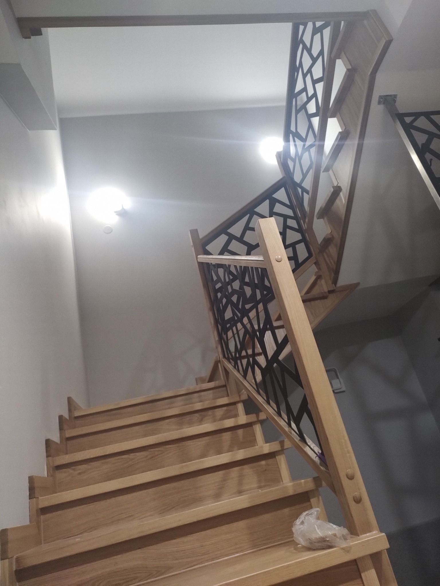 Drewniane schody z geometryczną, czarną balustradą, widok z dołu. Jasne drewno kontrastuje z ciemnym wzorem balustrady. Oświetlenie sufitowe.