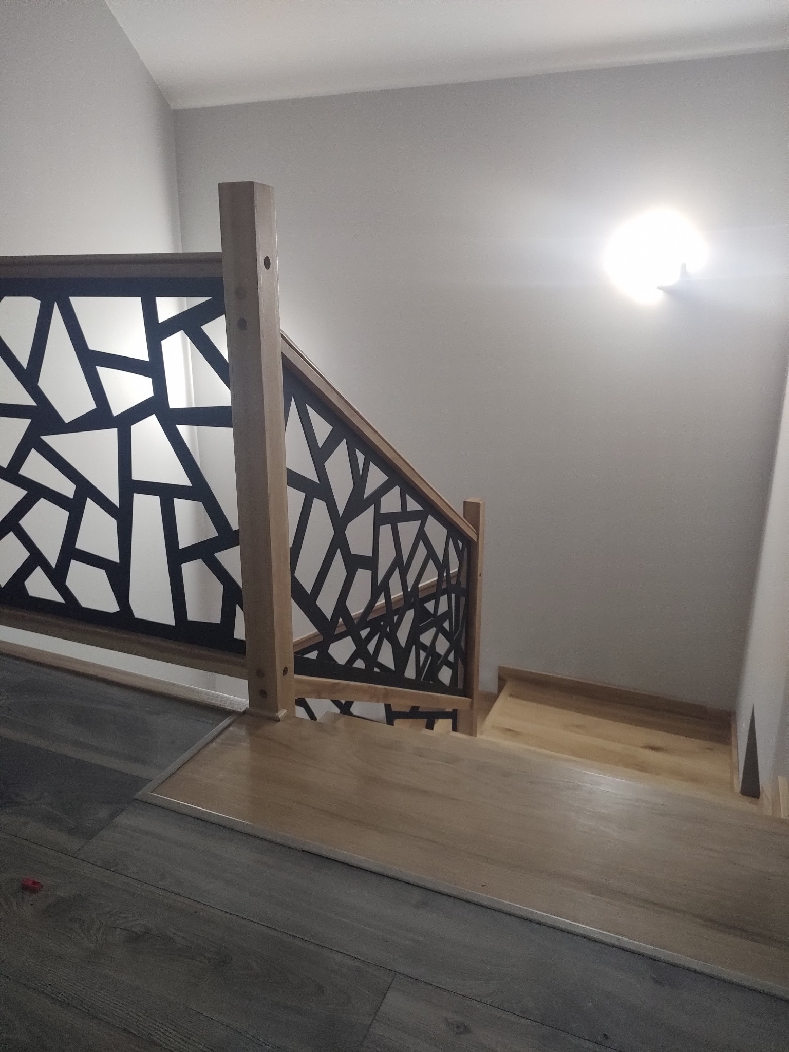 Drewniane schody z geometryczną, czarną balustradą. Widok z góry na pierwsze stopnie, łączące ciemną podłogę z jasnymi stopniami. Minimalistyczny design.