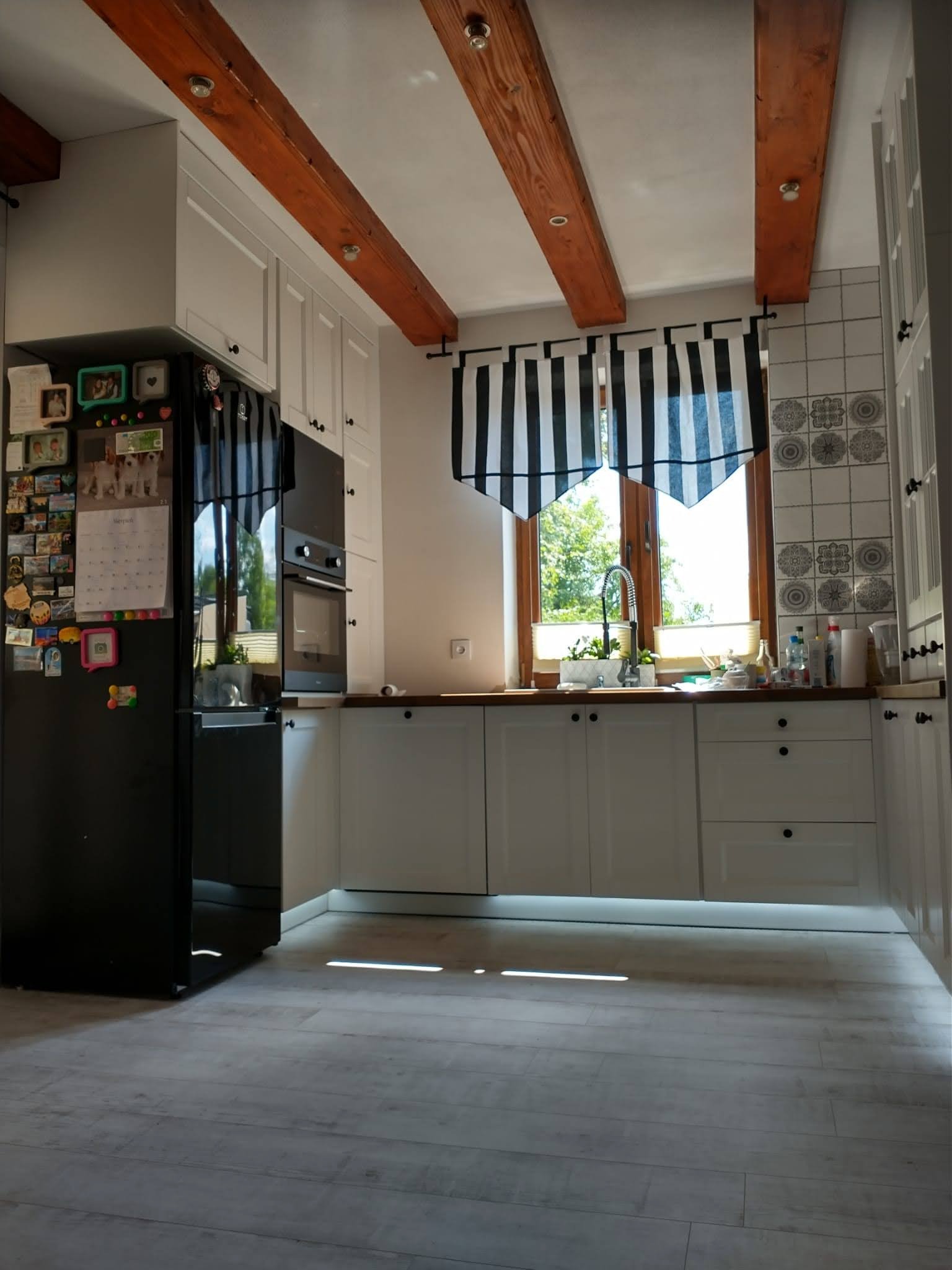 Jasna kuchnia z drewnianymi belkami stropowymi i czarną lodówką. Nad oknem krótka, biało-czarna, pasiakowa firanka. Na ścianie białe płytki z szarym wzorem.
