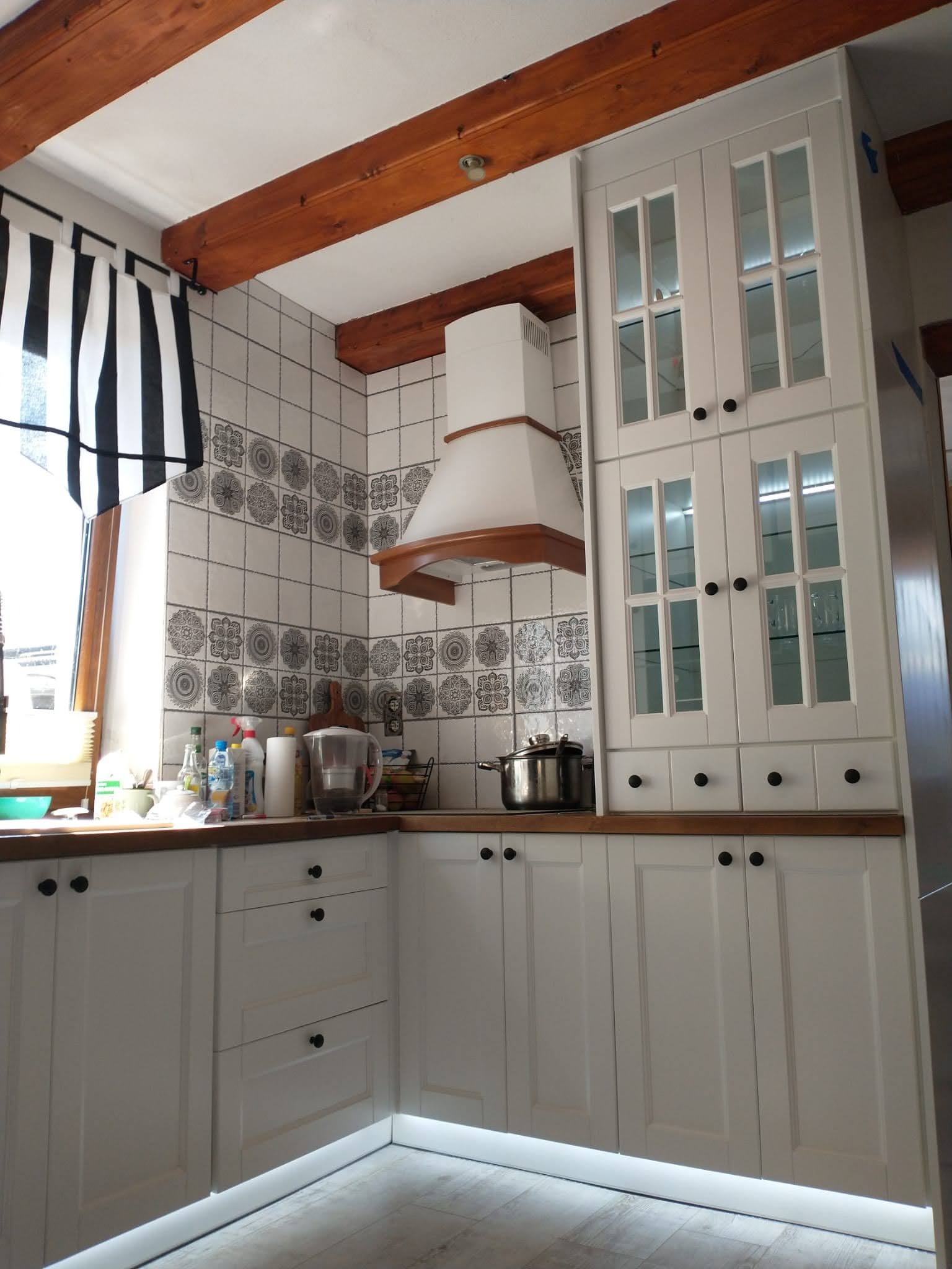 Biała kuchnia z drewnianymi belkami stropowymi i wzorzystymi płytkami. Okap podszafkowy i oświetlenie LED pod szafkami dodają nowoczesnego akcentu.