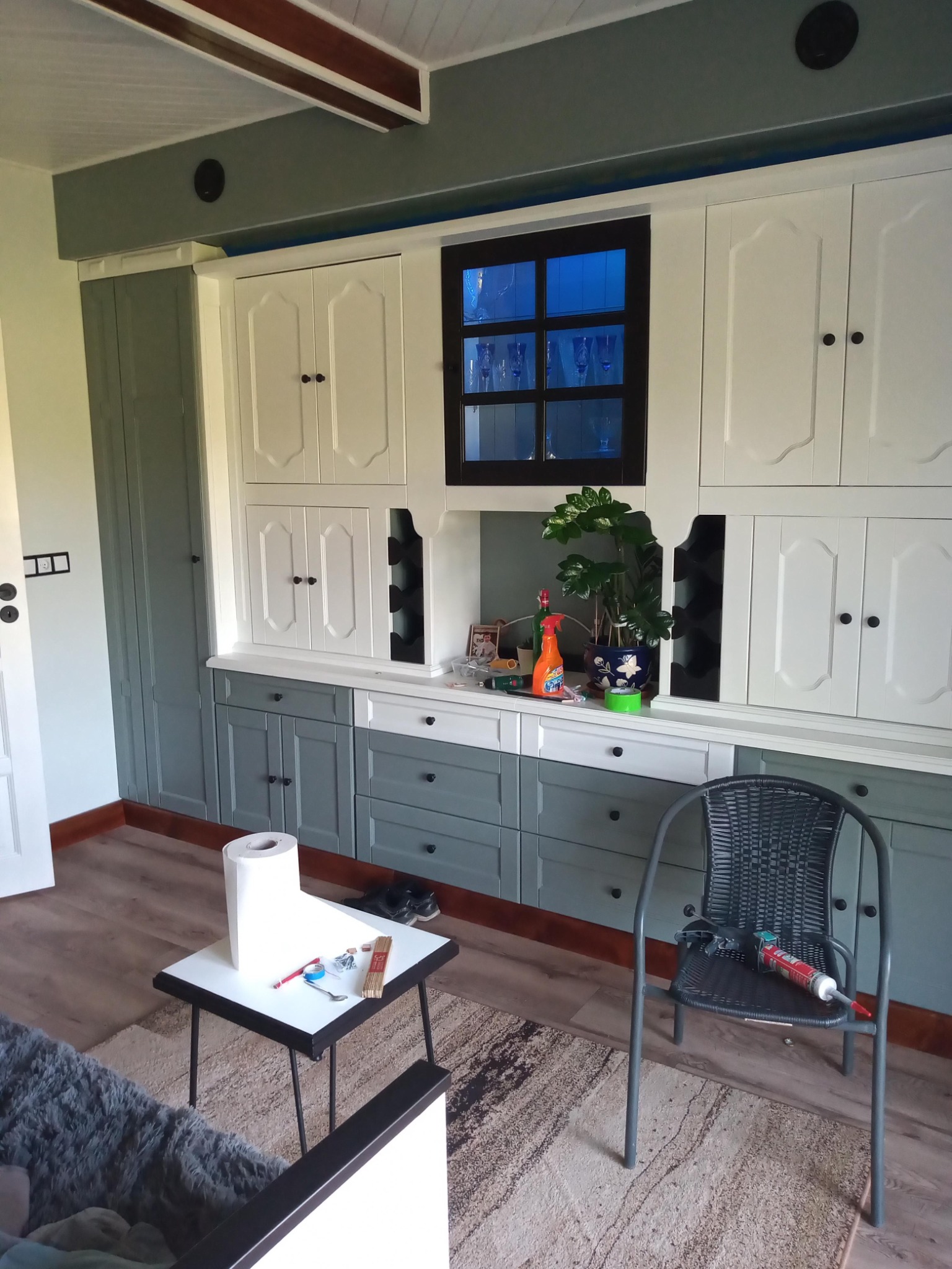 Wnętrze z zabudową meblową w odcieniach szarości i bieli, z widocznym oknem i elementami dekoracyjnymi. Na pierwszym planie stolik z narzędziami i krzesło z tubą kleju.