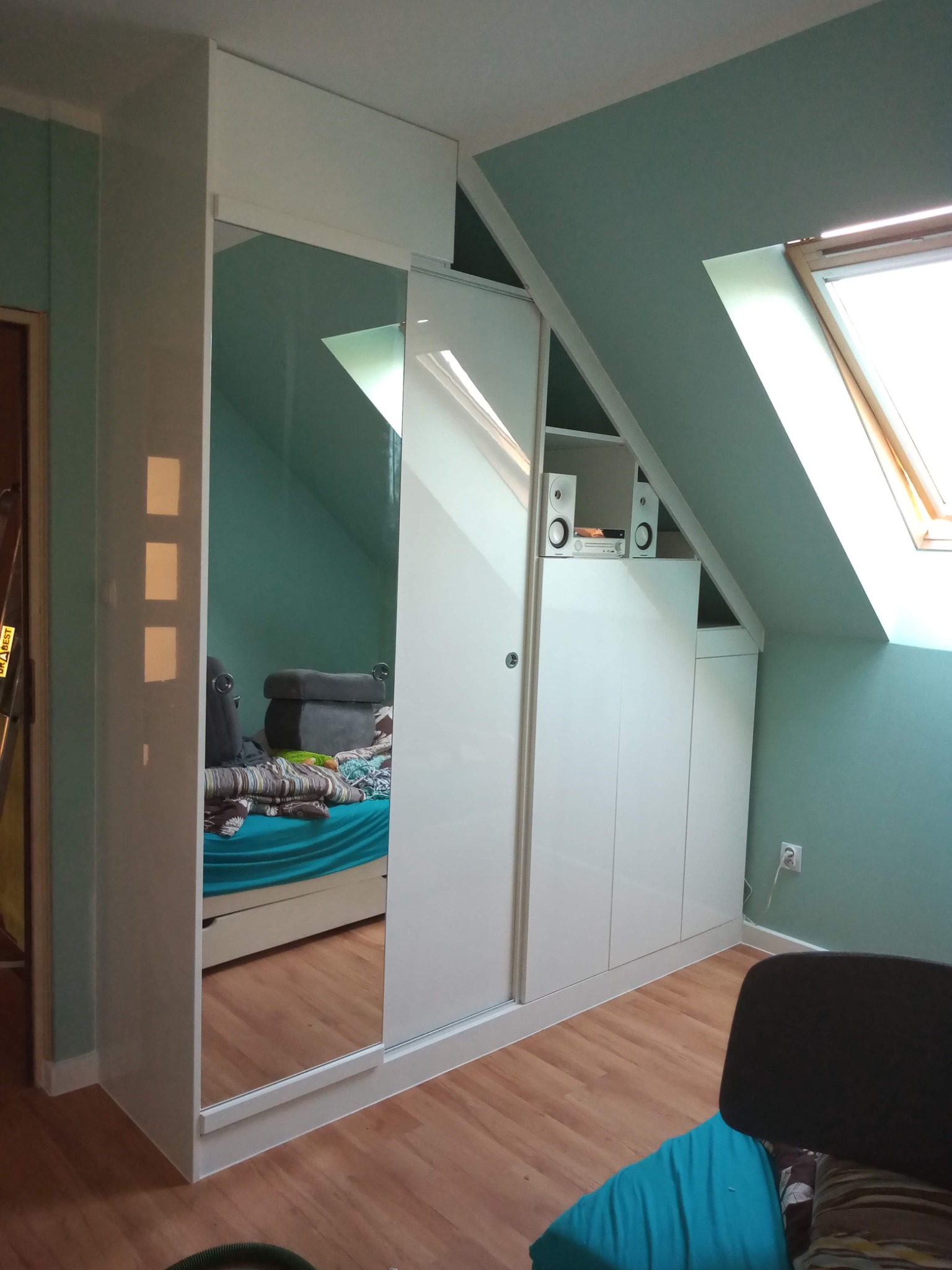 Biała szafa przesuwna z lustrem w pokoju na poddaszu, w tle okno dachowe i skośna ściana. Zabudowa meblowa pod skosem z dodatkowymi szafkami i półkami.