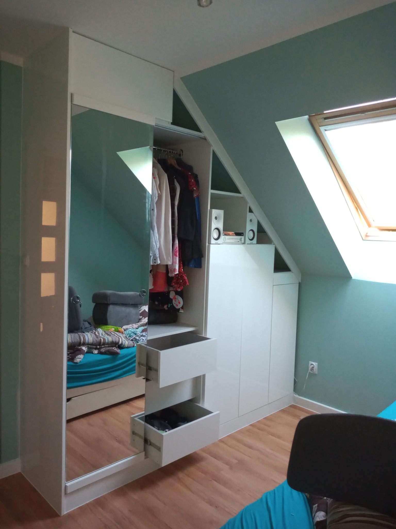 Biała szafa wnękowa z lustrem i szufladami, dopasowana do skosu poddasza. Widoczne ubrania wewnątrz i głośniki na półce. Jasne drewniane panele na podłodze.