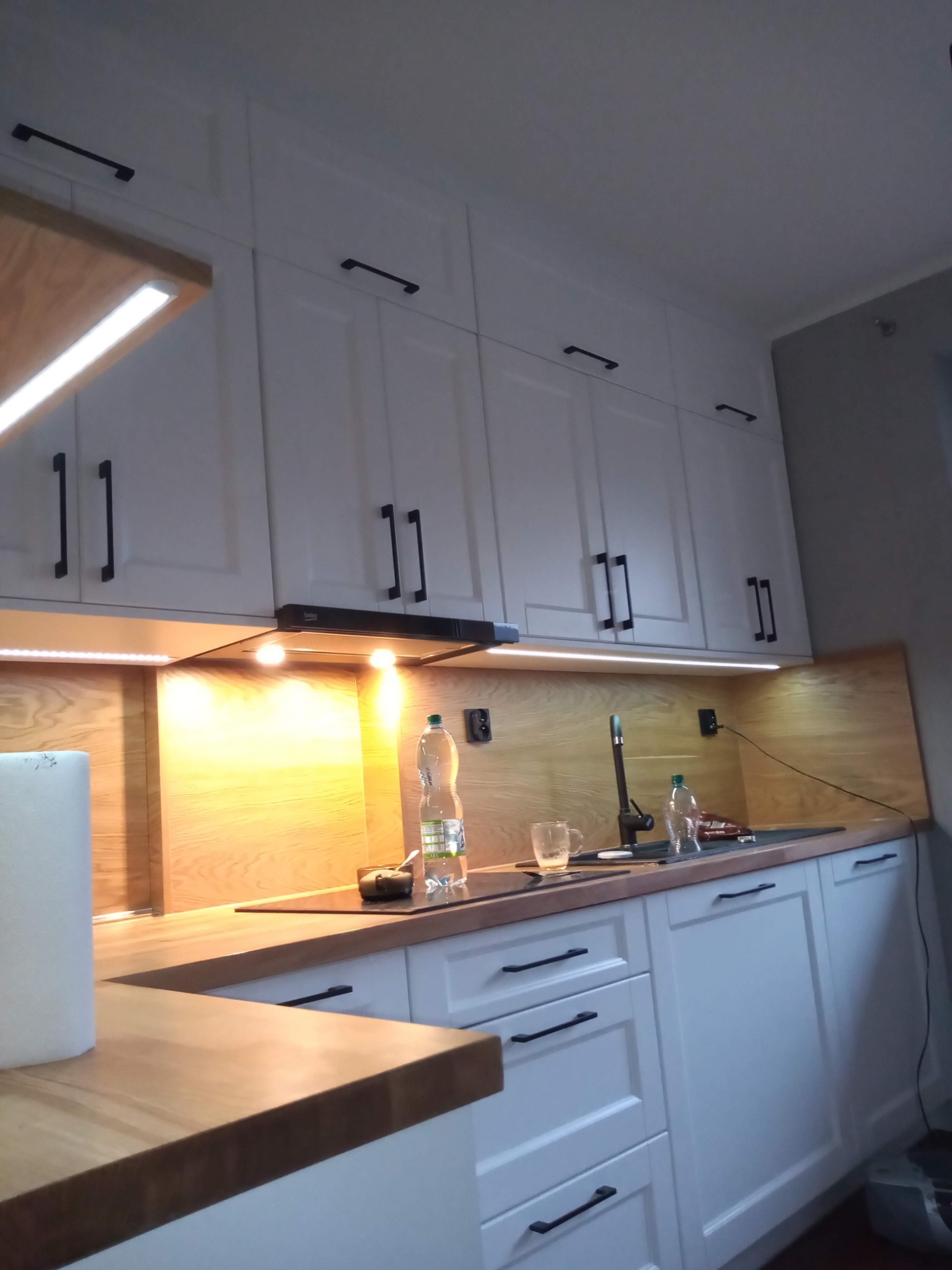 Biała zabudowa kuchenna z drewnianym blatem i czarnymi uchwytami, oświetlenie LED, Bulowice. Widok na zlewozmywak i górne szafki.