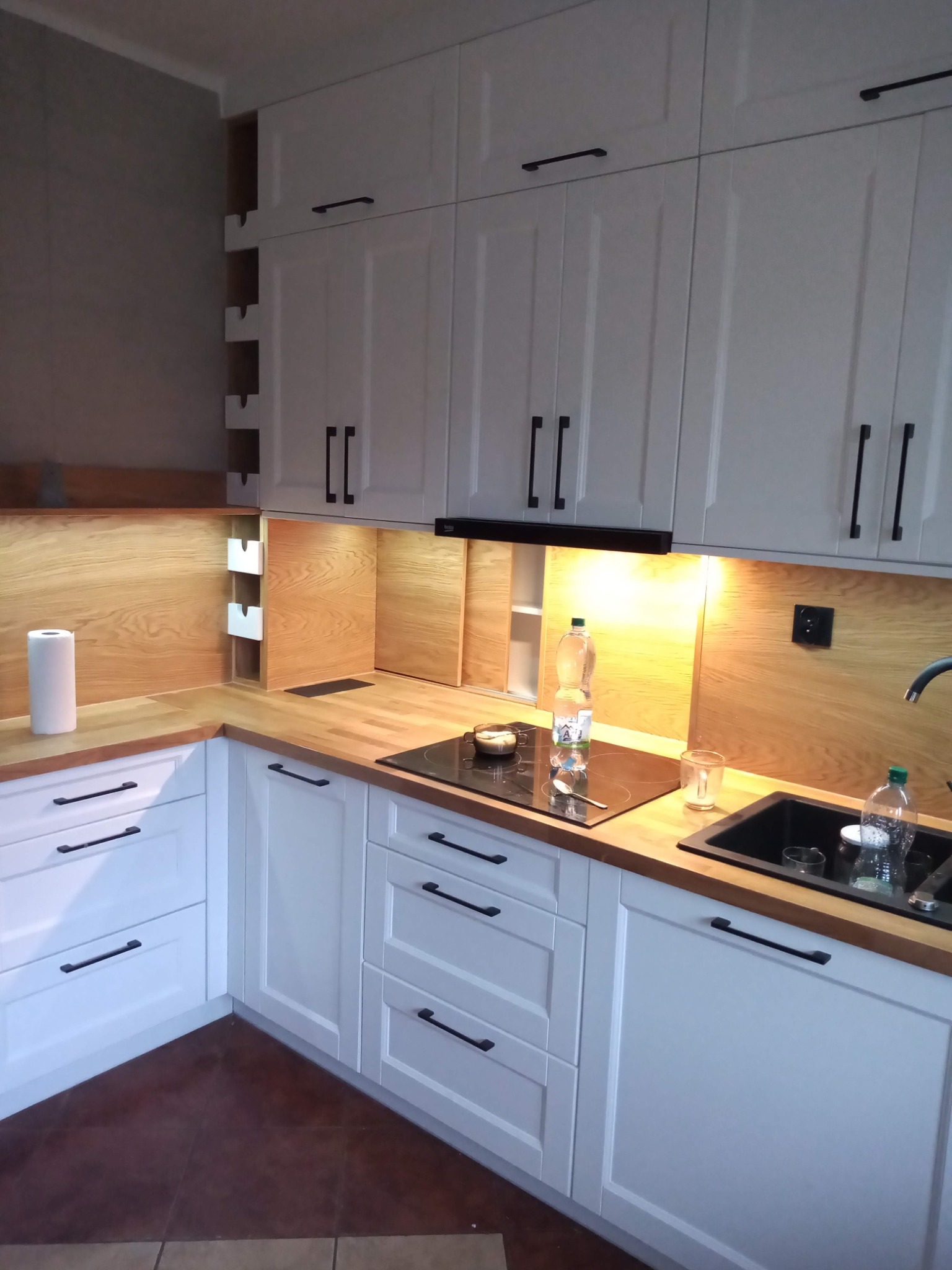 Biała kuchnia na wymiar z drewnianym blatem i czarnymi uchwytami. Widoczne szafki górne i dolne, płyta indukcyjna, zlew i fragment podłogi z brązowych płytek.